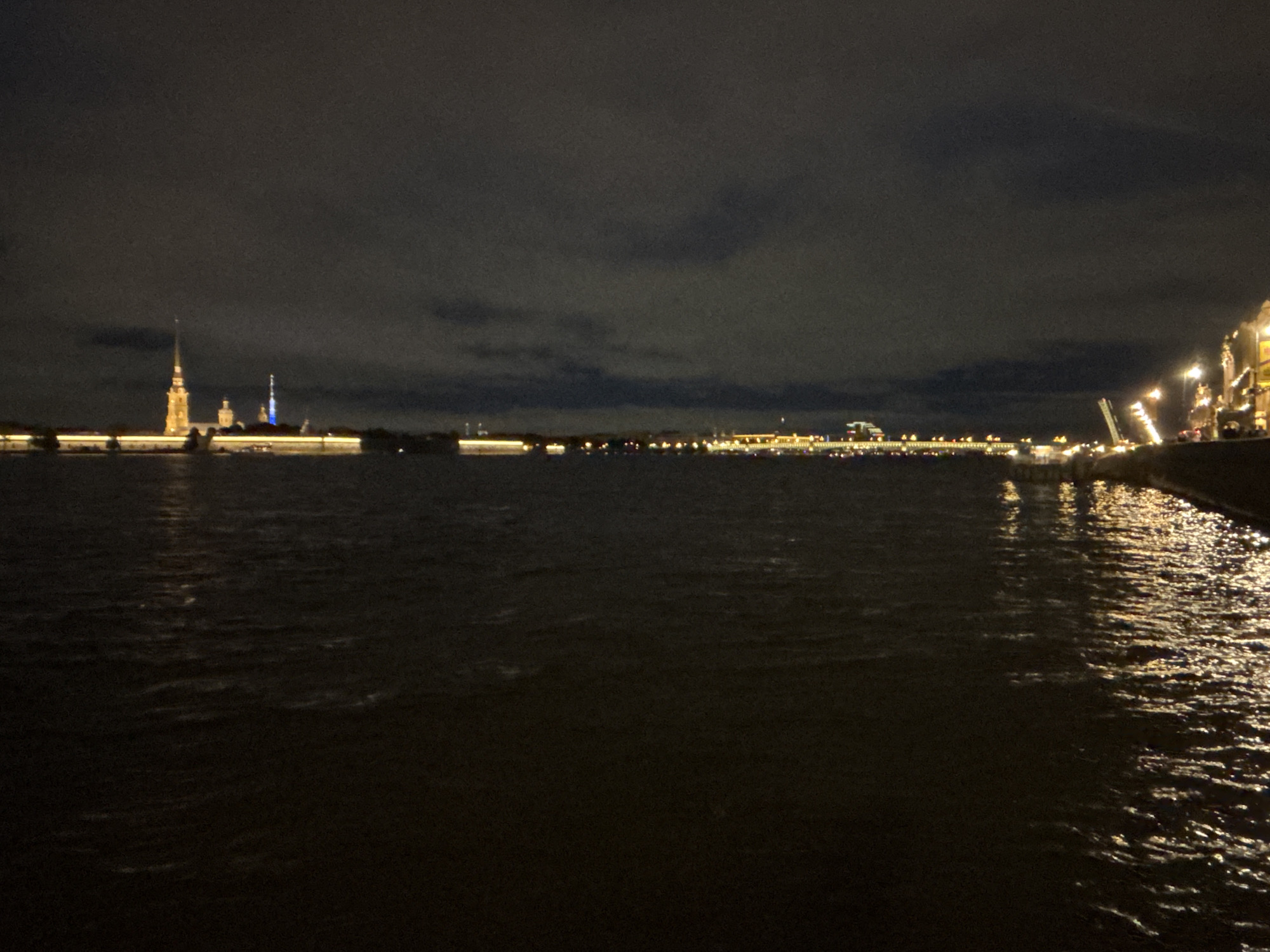The image shows a nighttime view of the Neva River in Saint Petersburg, Russia. In the background, the Peter and Paul Fortress is visible, illuminated with yellow and blue lights. Other illuminated buildings and bridges stretch along the horizon. The foreground is dominated by the dark, rippling water of the Neva River, reflecting some of the lights from the city. On the right, a section of the embankment is visible, lined with illuminated lampposts. The sky is overcast, contributing to the overall dark and moody atmosphere of the scene.