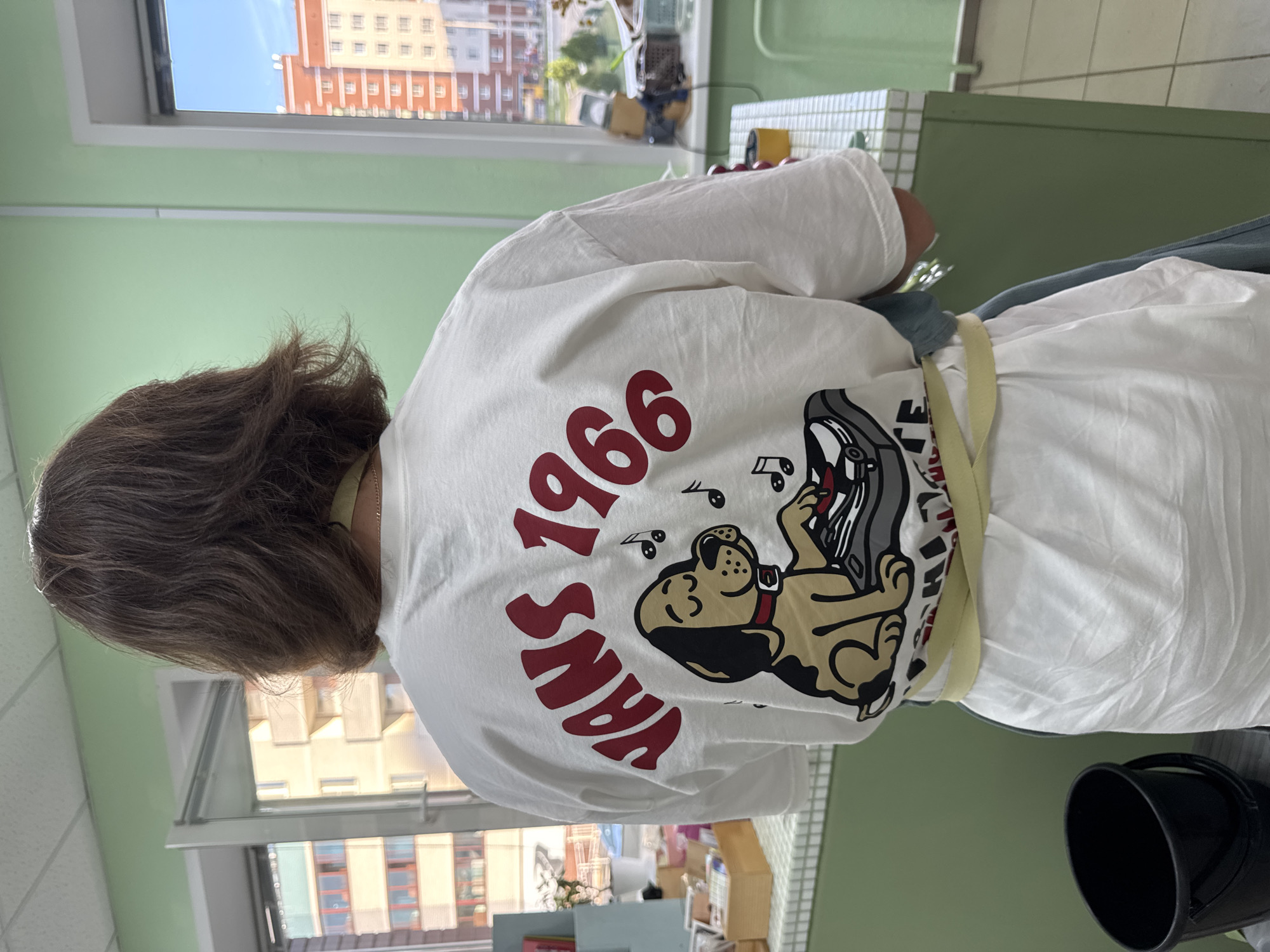 A woman with brown hair is wearing a white T-shirt with a "Vans 1966" design on the back, featuring a cartoon dog DJ. She has a light-colored apron tied around her waist. The scene is indoors, near a window with a view of buildings. Based on the architecture and appearance of the buildings visible through the window, the location is potentially Izhevsk, Russia. There is a black bucket on a counter in the foreground. The environment is indoors, and the room has light green walls.