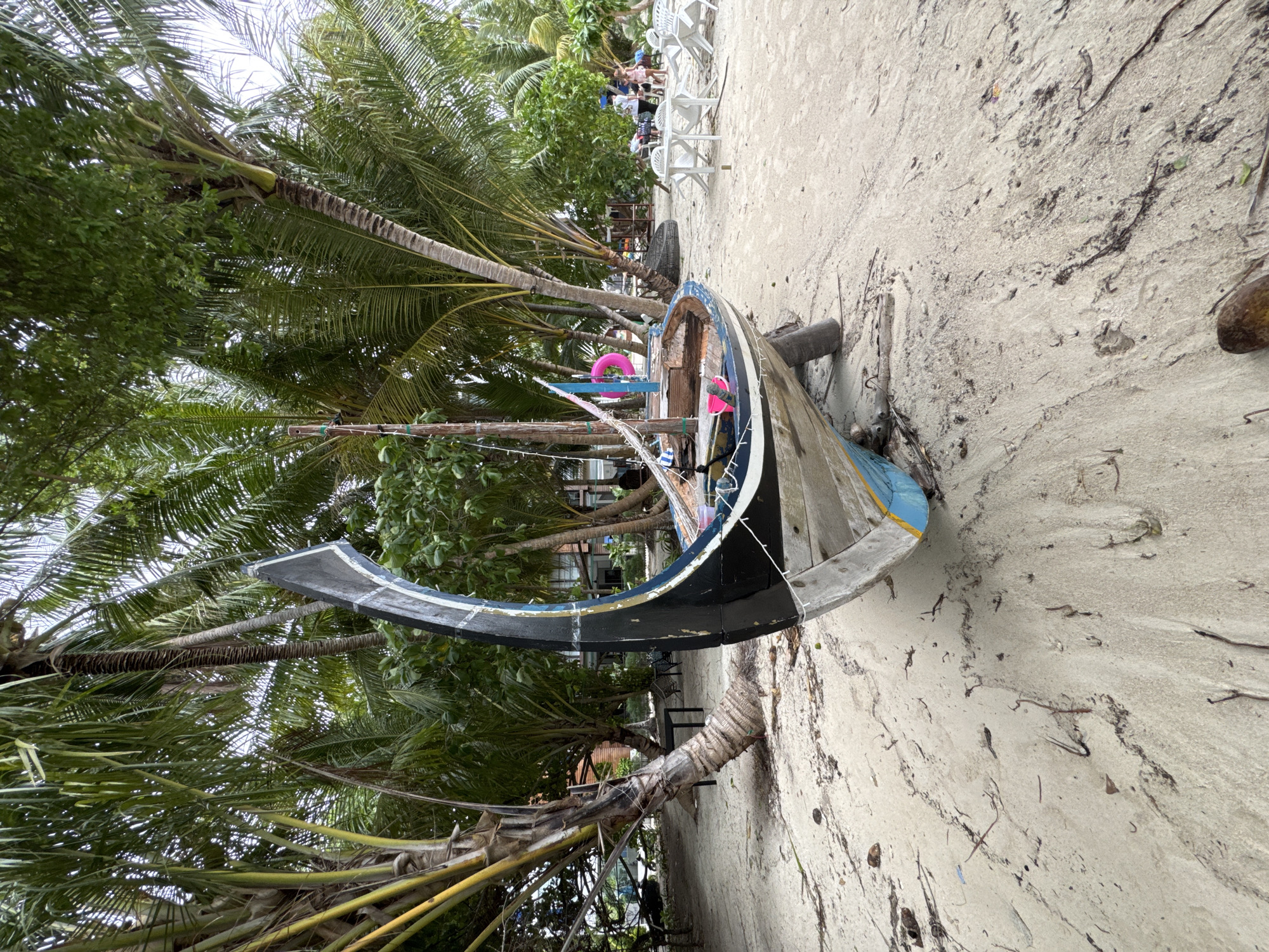 An abandoned wooden boat, with a high, curved black prow, rests on a sandy beach in Dhigurah, Maldives. The boat is positioned at an angle, revealing its weathered wooden hull with blue and white detailing along the sides. The interior of the boat contains a pink inflatable ring and a pink bucket. The beach is covered in light-colored sand with scattered seaweed and debris. Behind the boat, lush palm trees with long fronds create a dense canopy, casting shadows on the sand. In the background, a few people are visible standing near white plastic tables and chairs, suggesting a resort or dining area. The overall scene depicts a tropical beach setting with a prominent, aged vessel as the central focus.