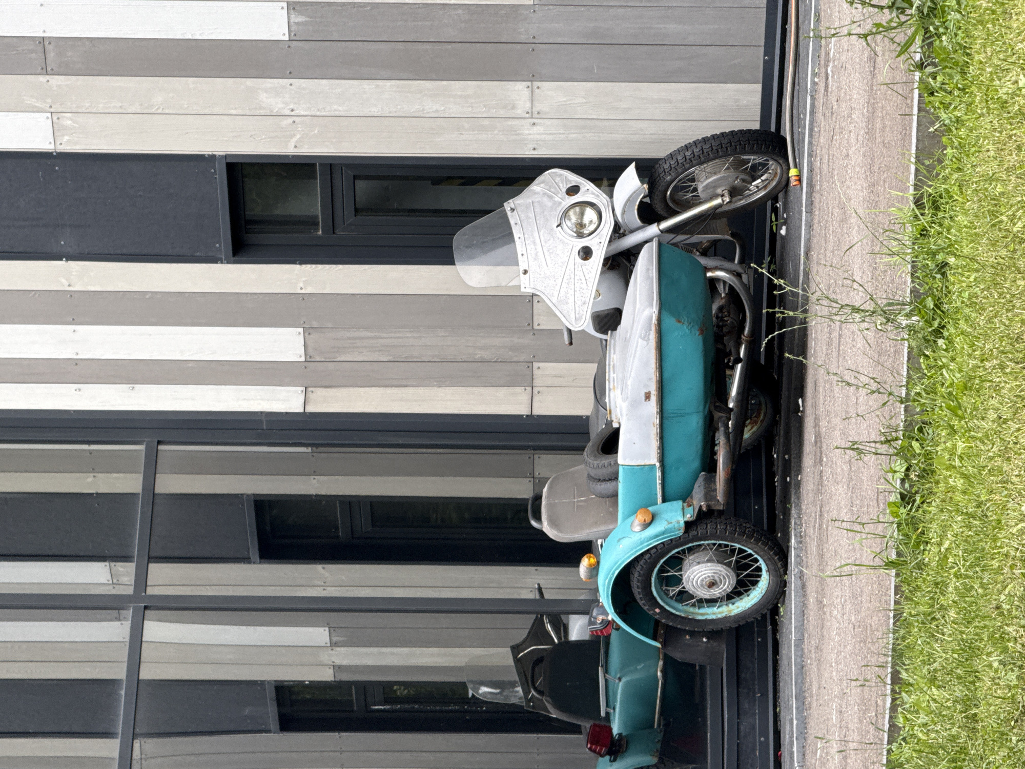 The image shows a three-wheeled motorcycle with a sidecar parked on an asphalt path next to a grassy area. The motorcycle is teal and white, with a large, silver windshield and a round headlight. It has two wheels in the back and one in the front. The background features a modern building with horizontal siding in shades of gray and white, and dark-tinted windows. The building appears to be multi-story, with a reflective facade that mirrors the surrounding environment. The grassy area on the right is lush and green, with some weeds growing taller than the grass. The lighting suggests it is daytime, possibly a sunny day, given the bright illumination of the scene. The image is oriented vertically, with the motorcycle appearing to be on its side relative to the viewer's orientation. No people are visible in the frame, and no text is present. The scene seems to be set in an urban or suburban environment, possibly a park or an area adjacent to a contemporary architectural structure. The motorcycle's design suggests it might be vintage or a custom build.
