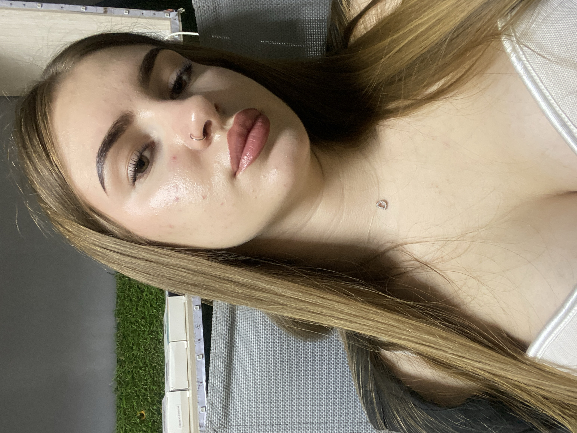 A young woman with long, brown hair is posing for a selfie. She is wearing a white top and a necklace with a small pendant. Her makeup includes foundation, lipstick, and mascara. She has a nose ring. In the background, there is a grey textured surface, possibly furniture, and a strip of artificial grass. There are also some white boxes stacked on a shelf with LED lights. The lighting suggests it is indoors or an outdoor covered area.