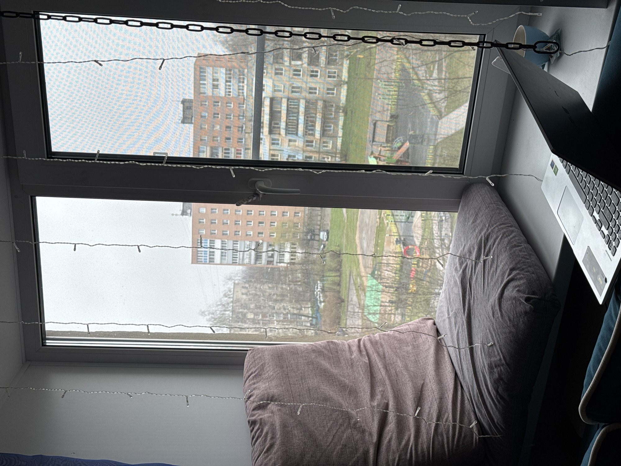 The image displays an indoor window view overlooking an urban landscape in Ново-Никольское, Russia. The window frame is dark, and string lights with small clear bulbs are strung horizontally across both upper and lower panes. A black decorative metal chain also spans the upper window section.
On the wide windowsill, a cozy arrangement of pillows creates a seating area; one pillow is a muted reddish-purple, and another is grey. To the right, a silver laptop is open, revealing its keyboard and trackpad, beside a light blue mug.
The outdoor scene features multiple multi-story apartment buildings with varied facades, including light concrete and reddish-brown brick, typical of the region. Bare trees are visible, suggesting late autumn, winter, or early spring. Patches of green grass or a park area are present, along with faint indications of a children's playground structure. The weather appears overcast and grey, providing diffused natural light.
No specific activity is explicitly depicted, but the setup suggests a comfortable, contemplative space for working or relaxing by the window. A small green sticker is visible on the laptop, but no other discernible text is present.
