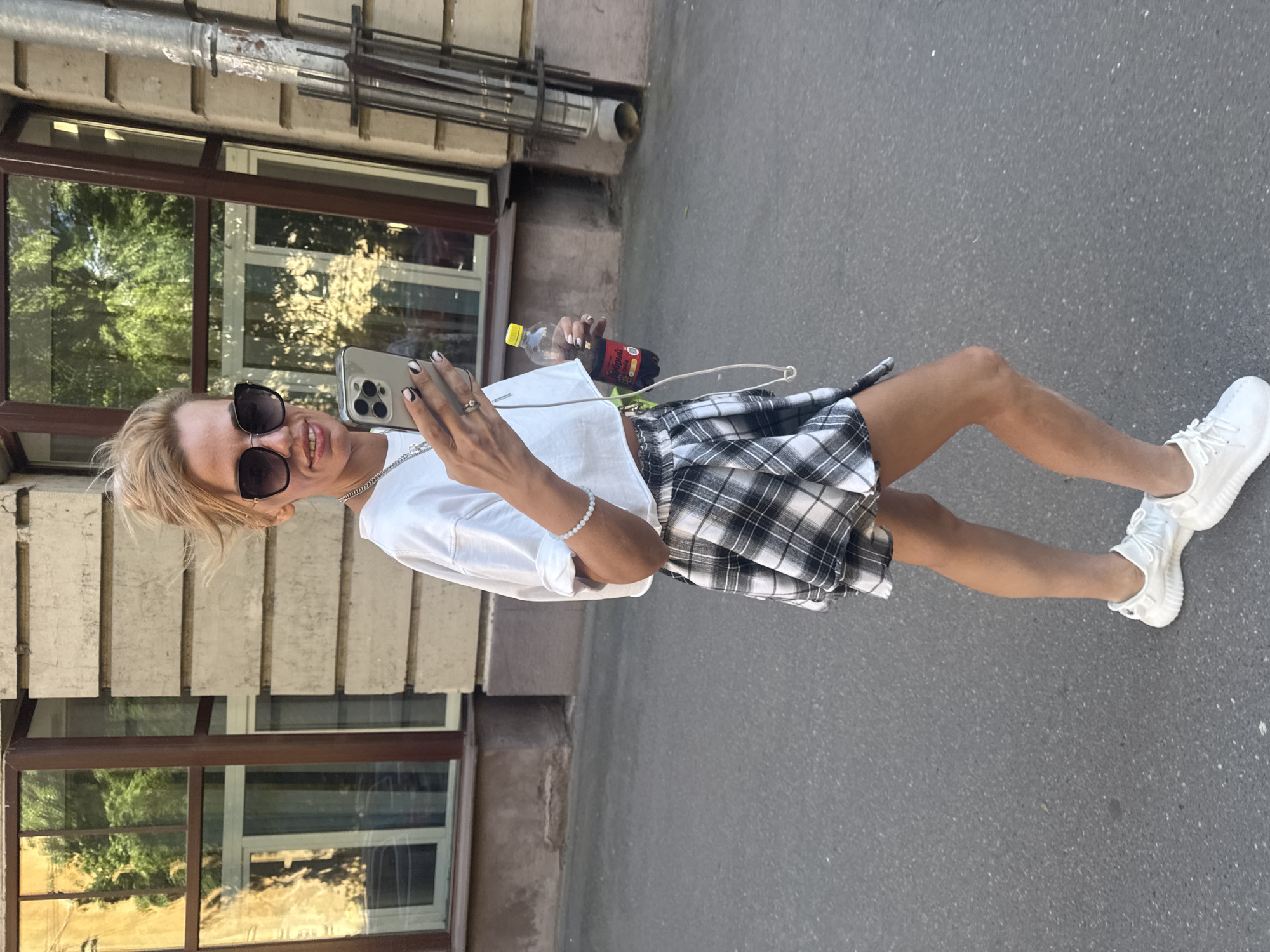 A blonde woman with sunglasses and a white crop top, black and white plaid skirt, and white sneakers walks on a sidewalk in Saint Petersburg, Russia, holding a phone and a bottle of soda. The background shows a building with windows and a drainpipe. The lighting suggests it is daytime, possibly late afternoon, with the sun casting shadows on the street.