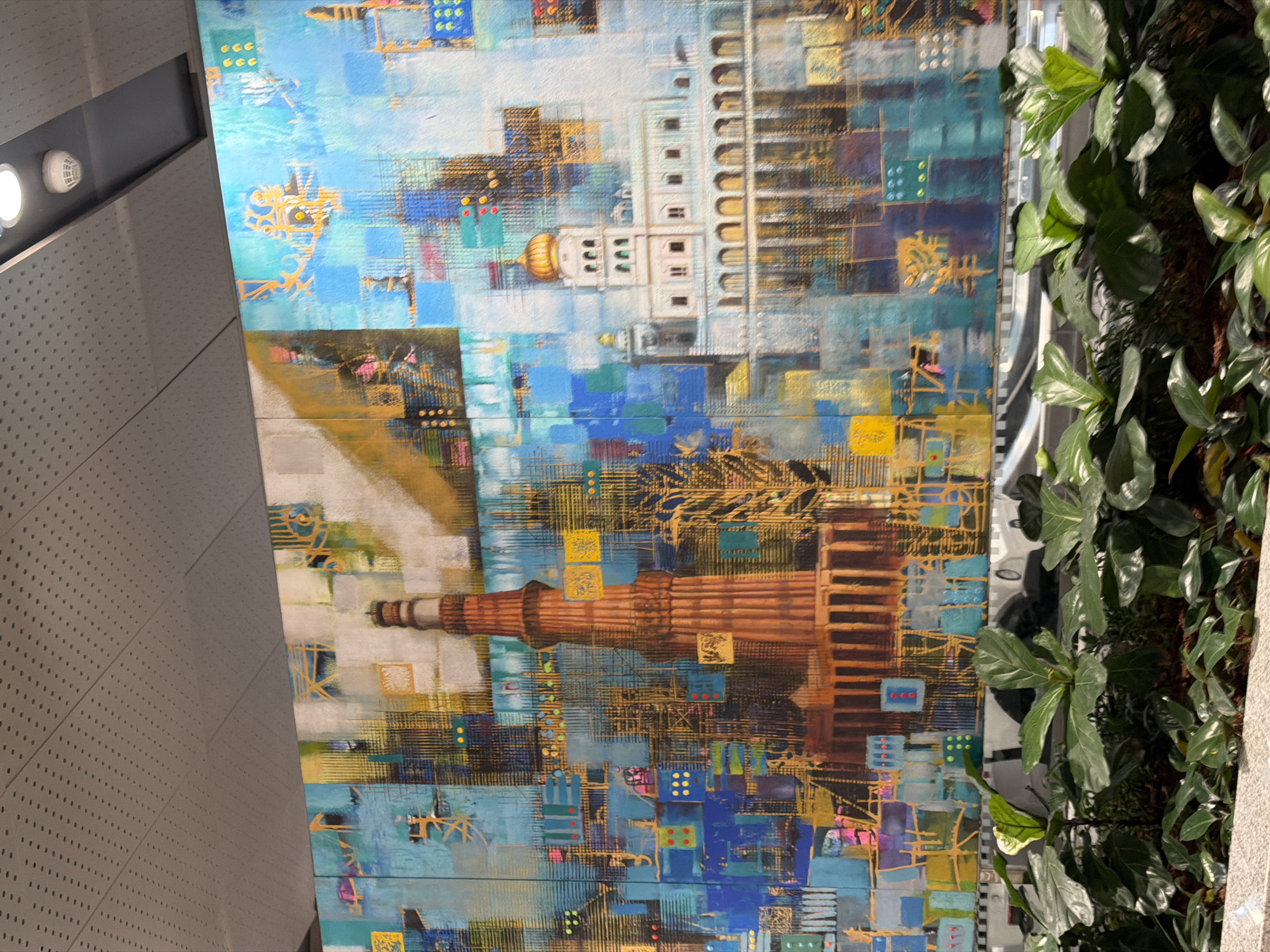 The image is a photograph of a large, colorful mural in a public indoor space. The mural depicts architectural landmarks of New Delhi, India, with a focus on the Qutub Minar, a towering brick minaret, and a white domed building resembling a Sikh Gurudwara. The artwork is abstract and modern, featuring geometric shapes, vibrant blue and yellow hues, and intricate golden patterns that suggest a cityscape or a digital representation.
In the foreground, a well-lit planter box contains lush green foliage, including plants with large, glossy leaves and smaller, darker green plants. The backdrop for the mural is a gray, perforated ceiling, likely part of a modern building's interior, with a black beam and a white circular smoke detector visible. The overall impression is a blend of contemporary art and historical representation within an urban environment.