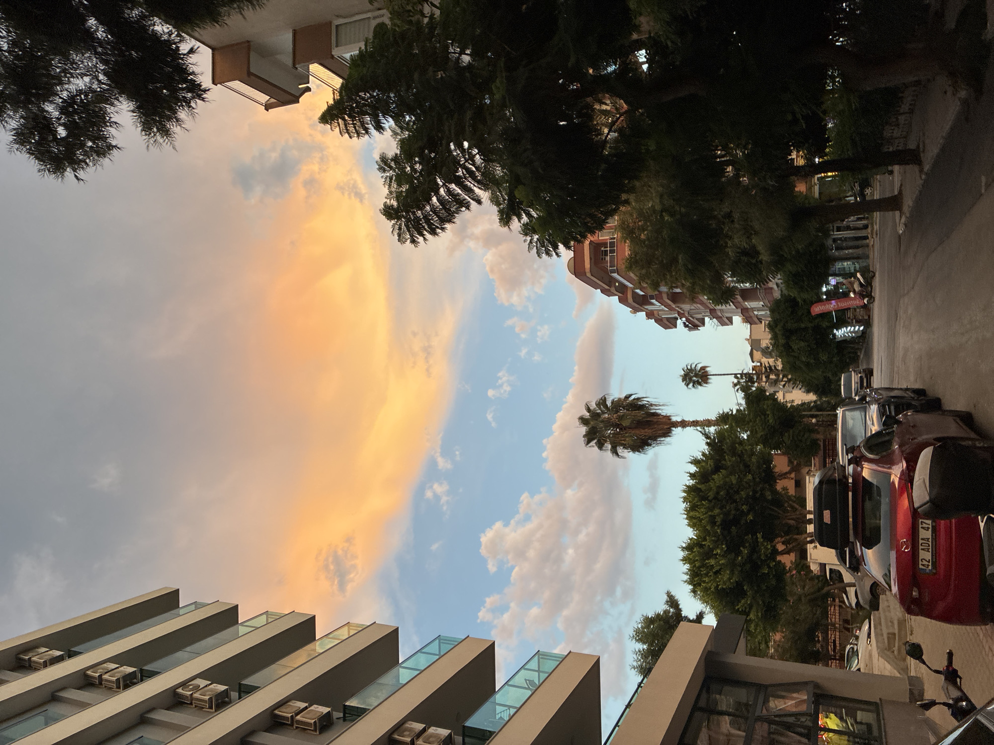 The image depicts a street scene in Alanya, Turkey, during sunset. In the foreground on the left, a modern building with multiple balconies, each equipped with air conditioning units, stands tall. Adjacent to it, a red sedan with a roof carrier and a license plate reading "42 ADA 47" is parked, along with other vehicles.
The street is lined with trees and buildings, some of which have visible balconies. A prominent palm tree stands in the middle ground. The sky above is a dramatic canvas of warm orange and yellow clouds, contrasting with the blue sky and scattered white cumulus clouds. The light suggests the end of the day, casting a warm glow on the scene. There are no people actively engaged in any discernible actions within the frame. The environment appears to be urban, with typical city infrastructure like parked cars and buildings.