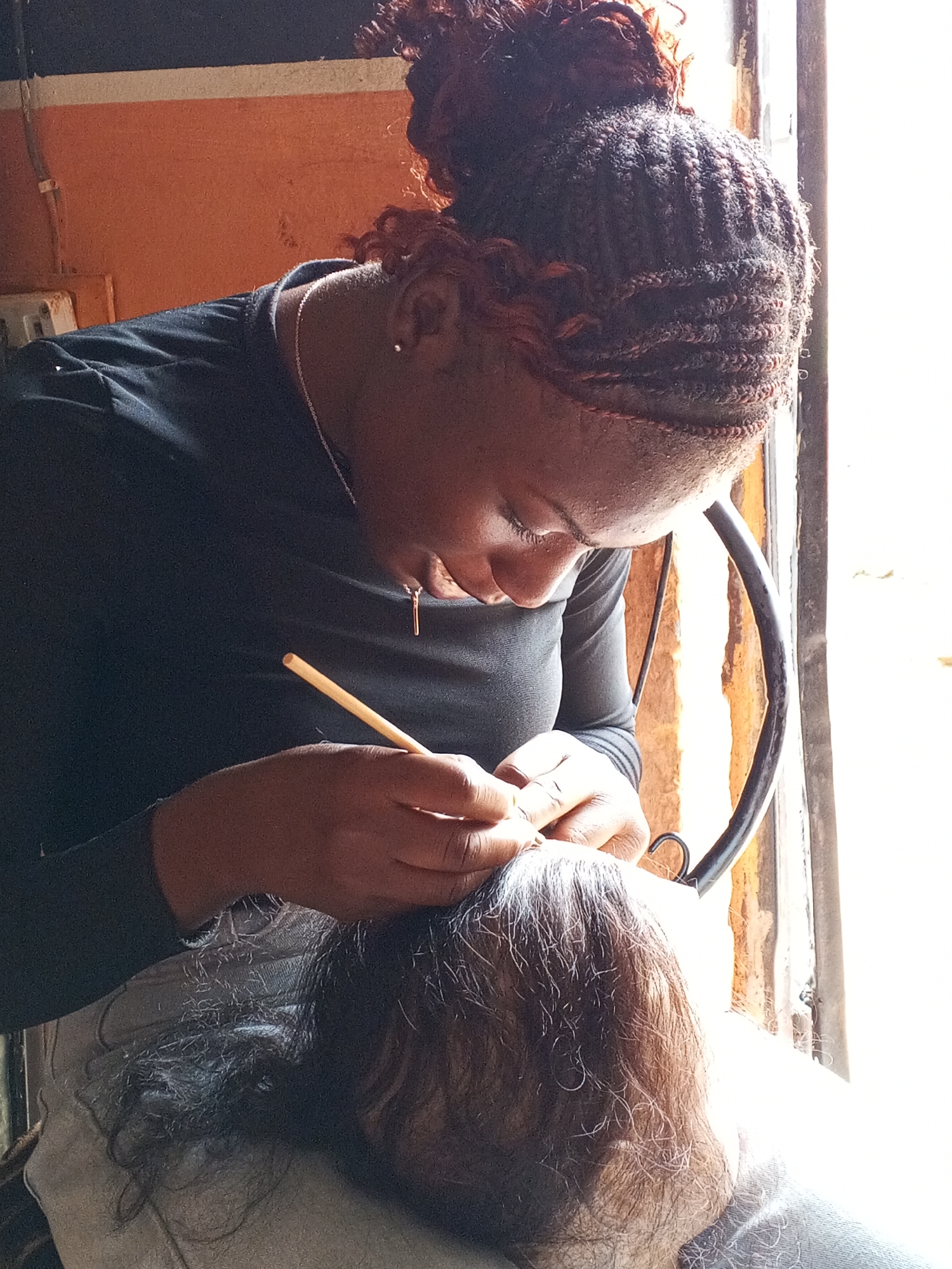 In a brightly lit interior location in Gwong, Nigeria, a woman with braided hair bunched on top of her head is focused on working on another person’s hair. She is wearing a dark, long-sleeved shirt and a delicate necklace with a pendant. She holds a light-colored, thin stick-like tool between her fingers as she works on the hair of the person below, who is seated and facing away from the camera. The hair of the person below appears gray and thinning. The setting appears to be near a window with a black metal frame. An orange wall is visible in the background.