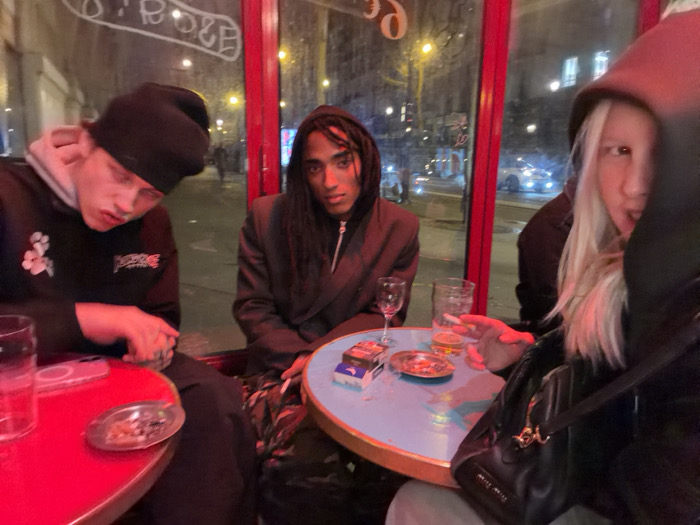 Three young adults are sitting at small tables inside a cafe booth. The person on the left wears a black beanie and hoodie, looking downwards. In the middle, a person with dreadlocks wears a blazer. The person on the right has light blonde hair and a dark hoodie. A blue table between the two on the right features cigarettes, ashtrays, and drinks. The table on the left is red. Streetlights and cars are visible through the window behind.