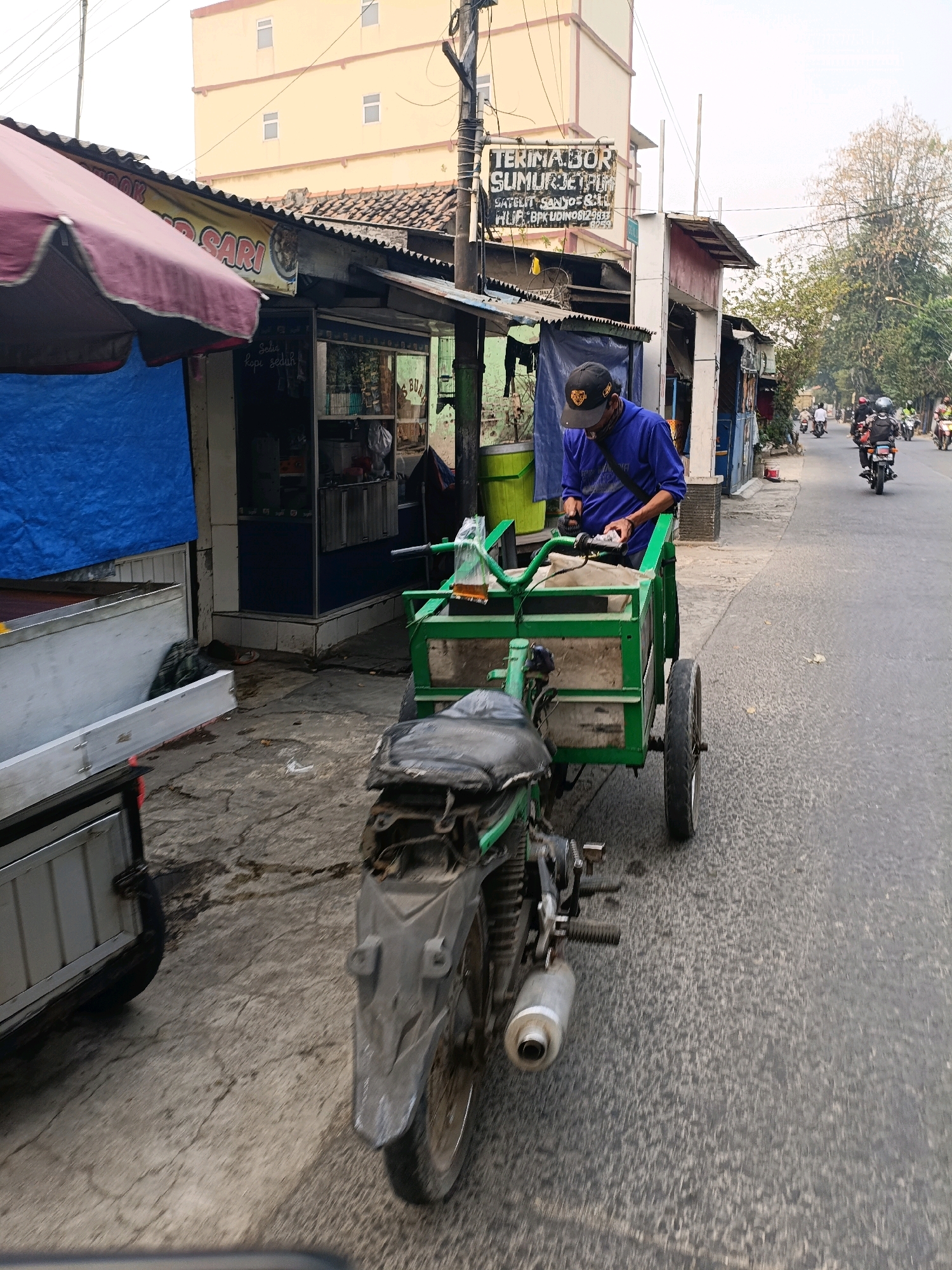 The image captures a street scene in Tangerang, Indonesia, on a moderately cloudy day. In the foreground, a person wearing a blue shirt and a black cap is seen operating a green motorized cart with a motorcycle engine. The cart is laden with items, and the person appears to be engaged in some task related to its contents. The cart itself is a utilitarian vehicle, likely used for transport or vending.
Behind the cart, a building with a yellow facade and tiled roof is visible, suggesting a commercial or residential area. Signs are posted on the buildings, one reading "TERIMA BOR SUMUR JET PUMP SATELIT - SANYO & DLL HUB: BPK UDIN 08128129833". This indicates services offered, specifically well drilling and pump installation.
Further down the street, other motorized vehicles, predominantly motorcycles, are seen moving in the same direction, indicating traffic flow. The road is asphalted, and the sidewalks are paved with concrete. The overall environment appears to be an urban street with a mix of commercial establishments and residential buildings. Trees and utility poles are also present, contributing to the urban landscape. The presence of a blue tarp covering a portion of a structure on the left side of the image suggests ongoing construction or a temporary covering. The scene conveys a sense of daily life and commerce in a bustling Indonesian city.