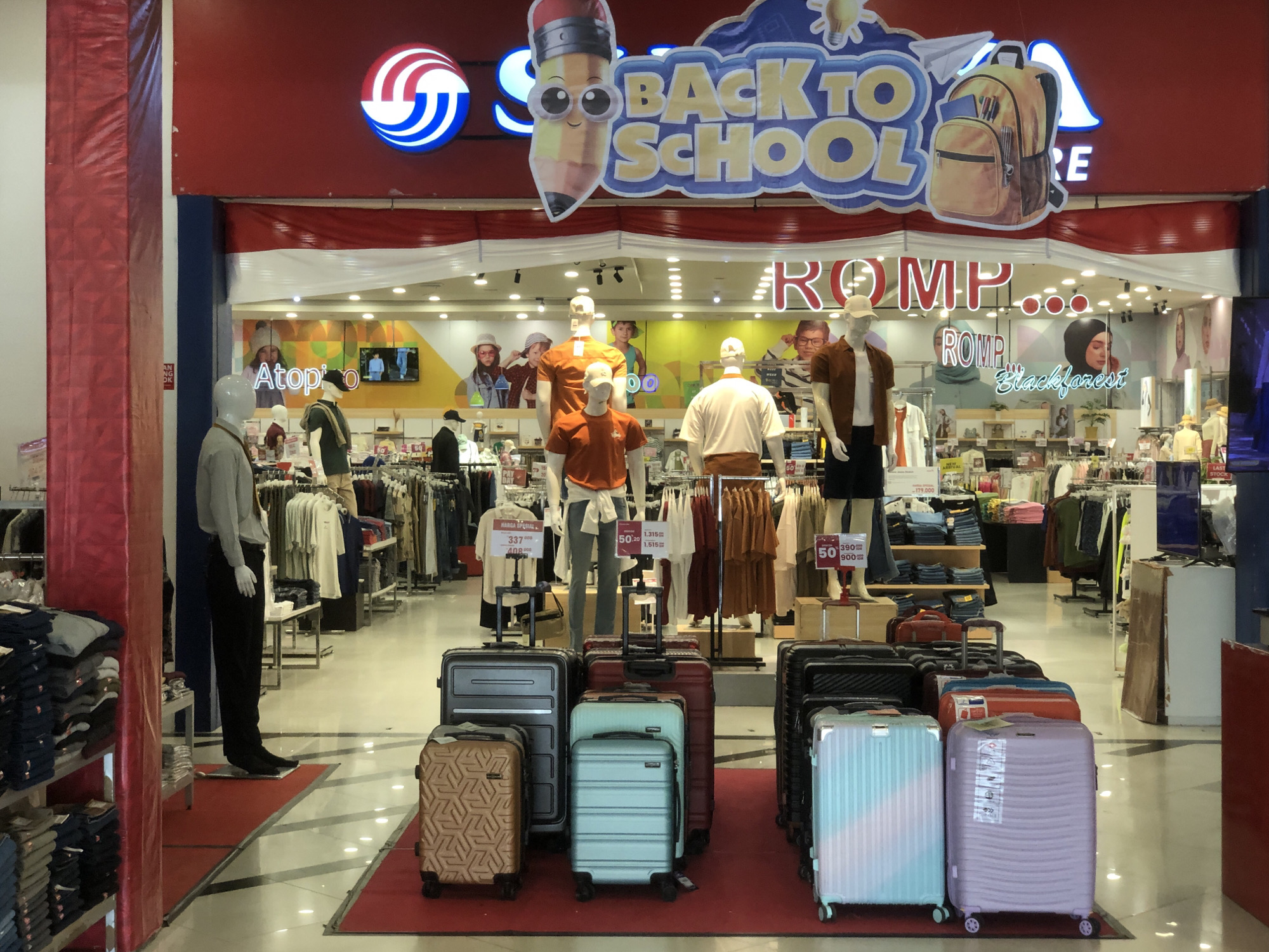 The image depicts the entrance of a store in Langsa, Indonesia, decorated for a "Back to School" promotion. The store's facade features a prominent red and white striped banner. Above the entrance, a large "Back to School" sign is displayed, complete with cartoon characters and a backpack graphic. Inside the store, mannequins are dressed in casual clothing, and racks of apparel are visible. A variety of luggage, including suitcases of different sizes and colors, are displayed in front of the store. The floor is tiled and reflects the overhead lighting. The overall scene suggests a retail environment preparing for a seasonal sale.