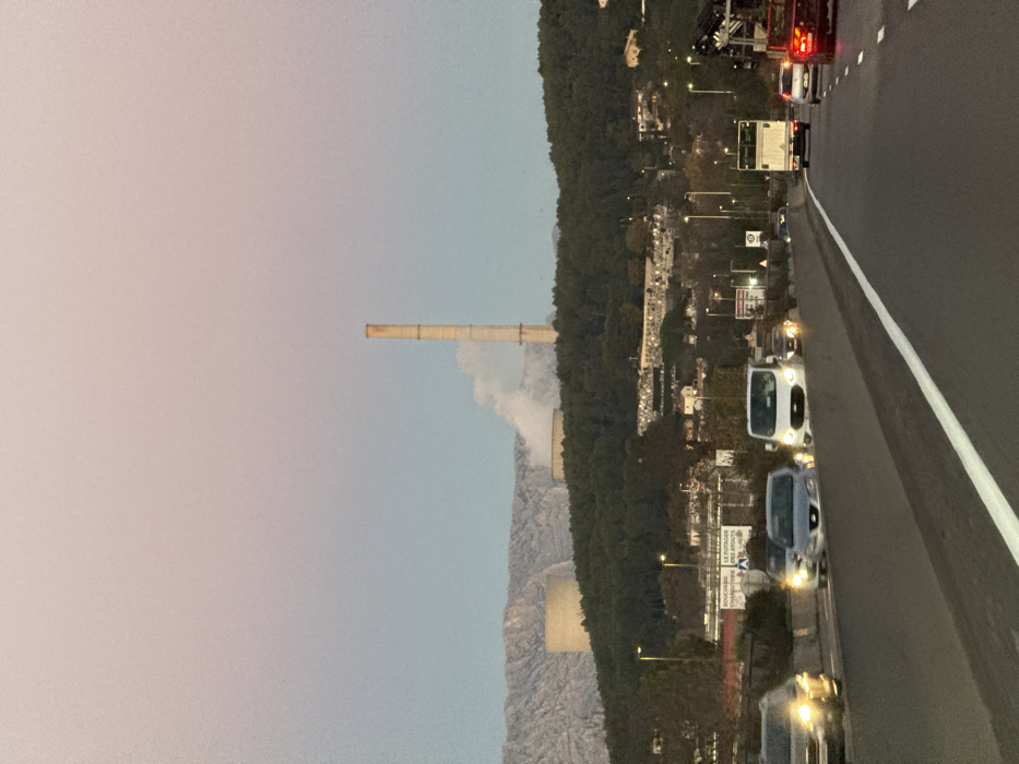 A tall chimney rises into the sky near a mountain range. White smoke billows from the chimney, partly obscuring the mountain. A highway with several vehicles runs parallel to a forest. Streetlights illuminate the scene, with advertisements and signs alongside the road. The sky is a gradient of colors, transitioning from a pinkish hue to a pale blue. The entire image is oriented sideways.
