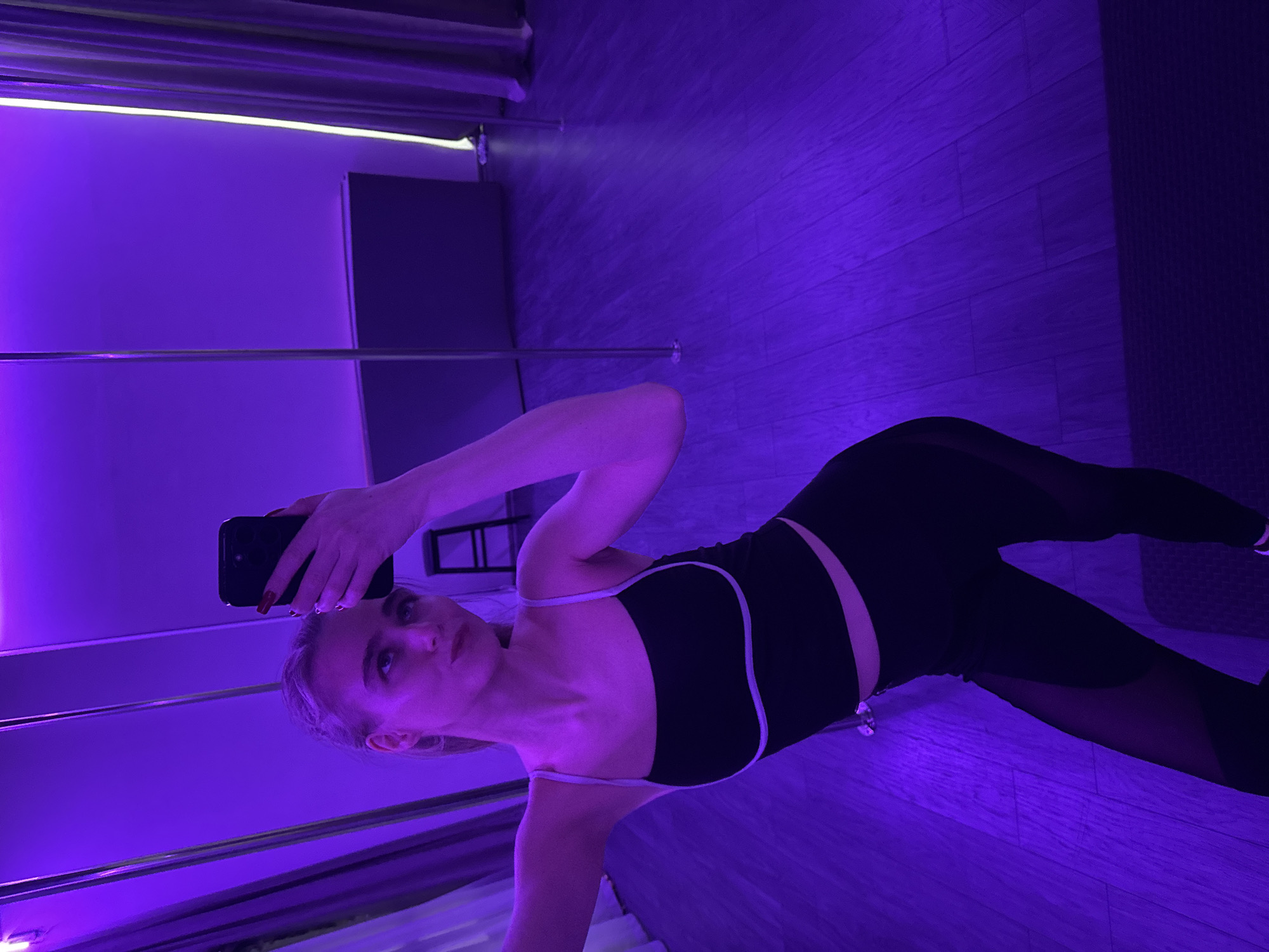 A woman is in a room with purple lighting. She is holding a smartphone and taking a selfie. She is wearing a black sports bra with white straps and black leggings with mesh inserts. The room has wood-like flooring. There is a black shelf behind her and a metal pole attached to the floor and ceiling on the left side. The location is in the city of Unknown, Unknown.