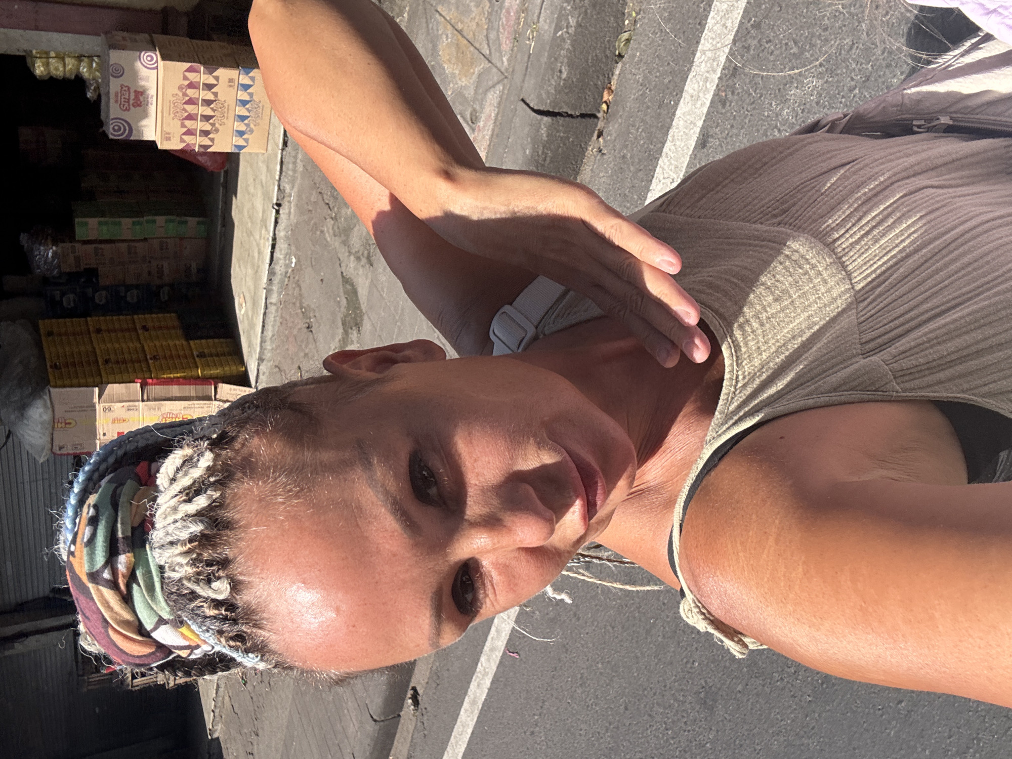 A light-skinned female stands outdoors on a paved surface in Kuta Selatan, Indonesia, with a storefront visible to her left featuring stacks of packaged goods. She is wearing a tan top and a colorful headscarf with braided hair visible at the front. The woman is holding one hand near her neck, and she appears to be taking a selfie. The backdrop includes a street with a painted white line.