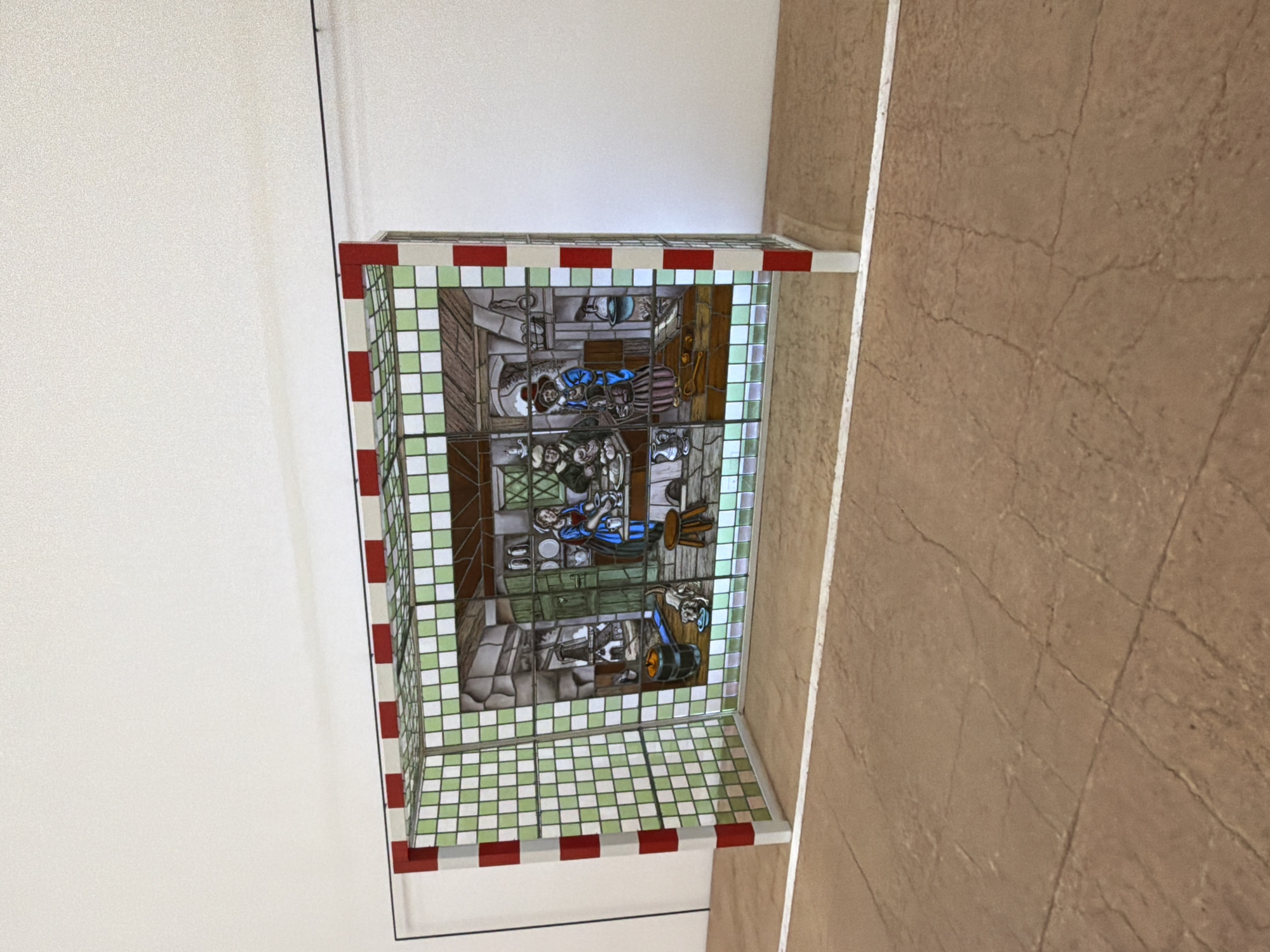 This image shows a stained-glass handball goal with a depiction of a historical domestic scene. The goal structure is made of white and green checkered panels, with red and white striped vertical supports. The stained glass within the goal shows an interior scene, possibly a kitchen, with several figures in period clothing. One figure, wearing a blue dress and white cap, appears to be preparing food at a table. Another figure, in a brown and blue outfit with a red hat, is standing near the table. A dog is visible in the lower left of the stained glass, near a barrel. The scene appears to be set in a dimly lit room with shelves of pottery and other kitchen items. The goal is placed on a brown, textured floor with faint lines, possibly indicating an indoor court. The background is a plain white wall. There is no indication of the location being in Mexico City, Mexico.