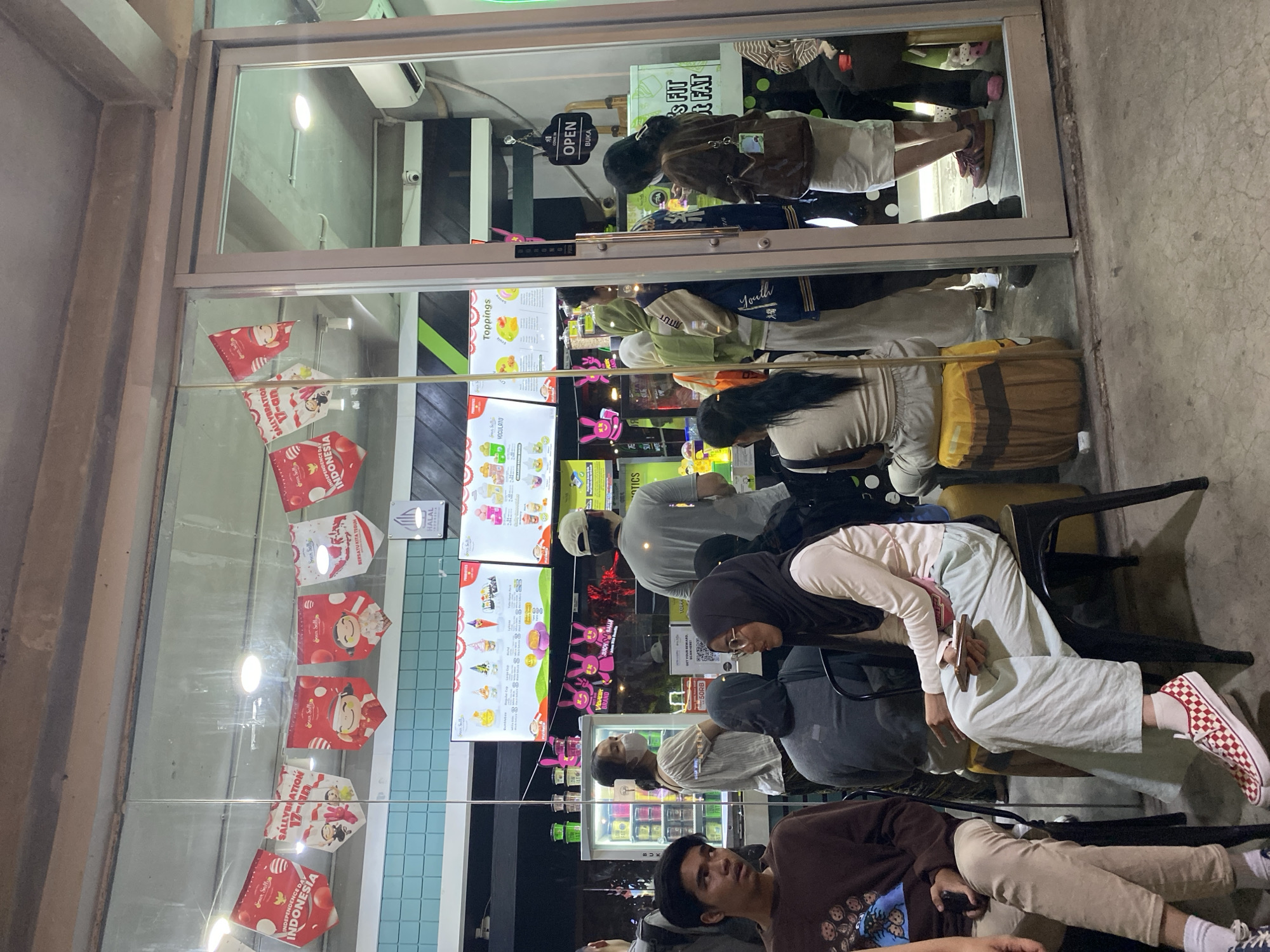 This image depicts the exterior of a food establishment, likely an ice cream or dessert shop, located in South Jakarta, Indonesia. The shop features a large glass storefront with an accompanying glass door. Inside, a brightly lit menu board displays various ice cream options and toppings. Red and white decorative banners, featuring Indonesian flag colors and possibly celebrating Independence Day, hang across the top of the storefront. A "HALAL" sign is visible.
Several people are visible, some queuing outside the shop and others inside. A man in a brown sweater and beige pants is seated on a chair on the left. A woman wearing a white top and a face mask is standing near a refrigerator. Another woman in a grey hooded garment is seated. In the center, a woman in a hijab and a light-colored top, along with a woman in a cream-colored top and light blue pants, are seated. Another group of people, mostly women in hijabs, are standing and moving inside the shop. The floor outside the establishment is paved concrete, and the surrounding environment is not clearly discernible. The overall atmosphere suggests a busy evening at a popular local eatery.