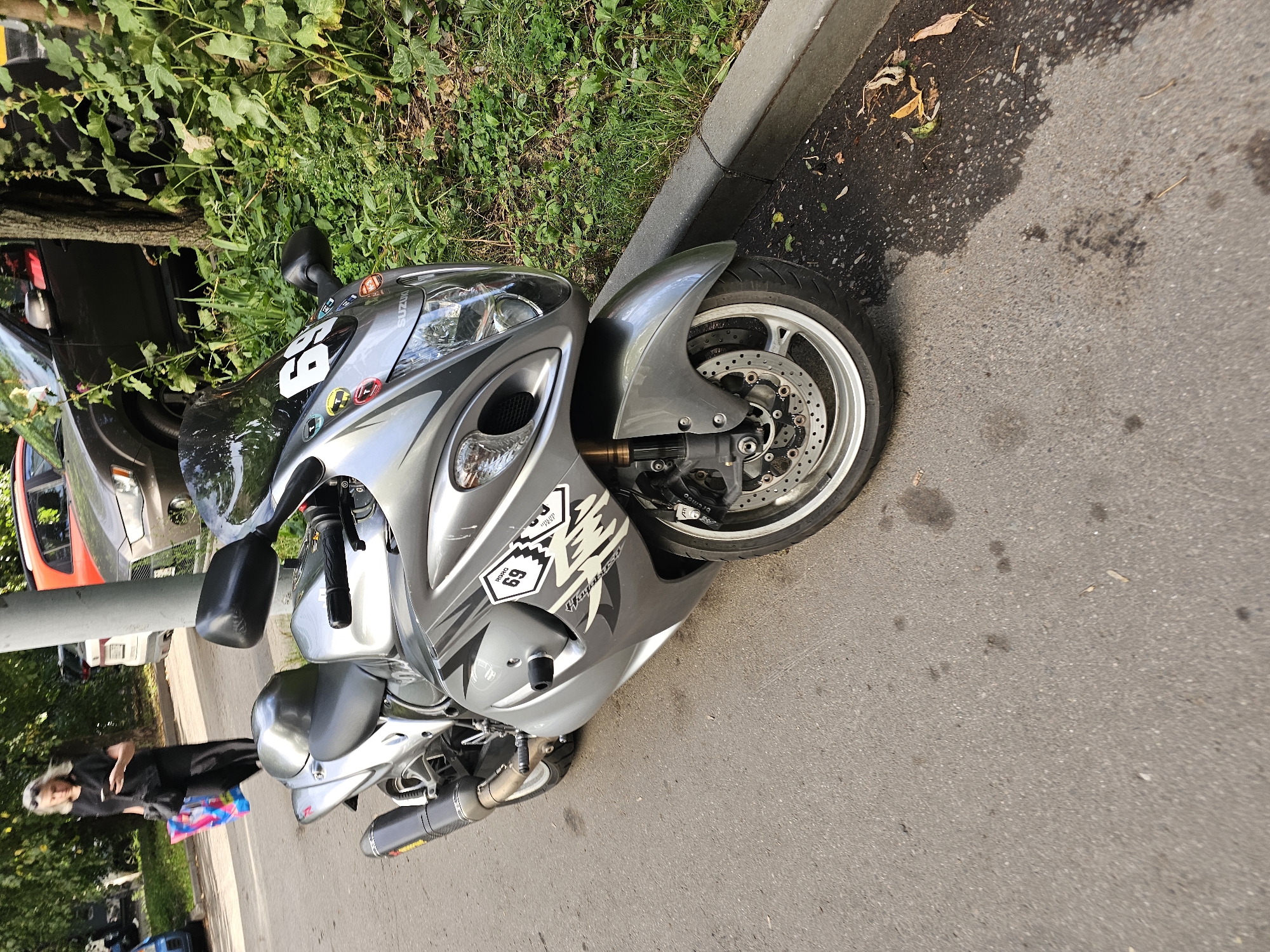 A silver Suzuki Hayabusa motorcycle is parked on the side of an asphalt road next to a curb. A woman in black clothing is walking away from the motorcycle and towards the left of the frame, carrying a colorful shopping bag. In the background, there are several cars and lush green vegetation. The scene appears to be outdoors in a city environment.