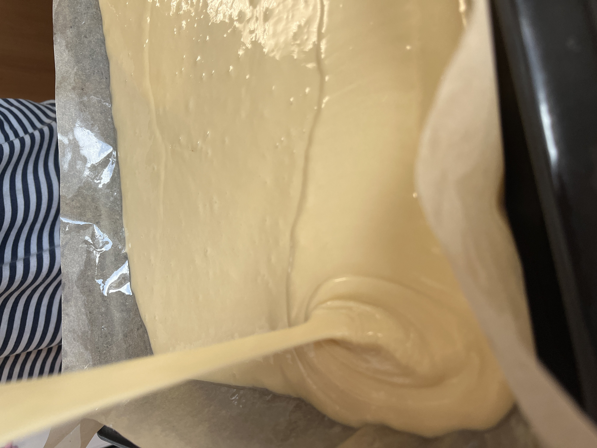 A close-up shot captures a stream of pale yellow batter being poured into a baking pan lined with parchment paper. The batter is thick and smooth, creating ripples as it settles. A person wearing a striped shirt is partially visible in the upper background. The scene appears to be in a kitchen, likely in Chaikovsky, Russia, based on the query.
