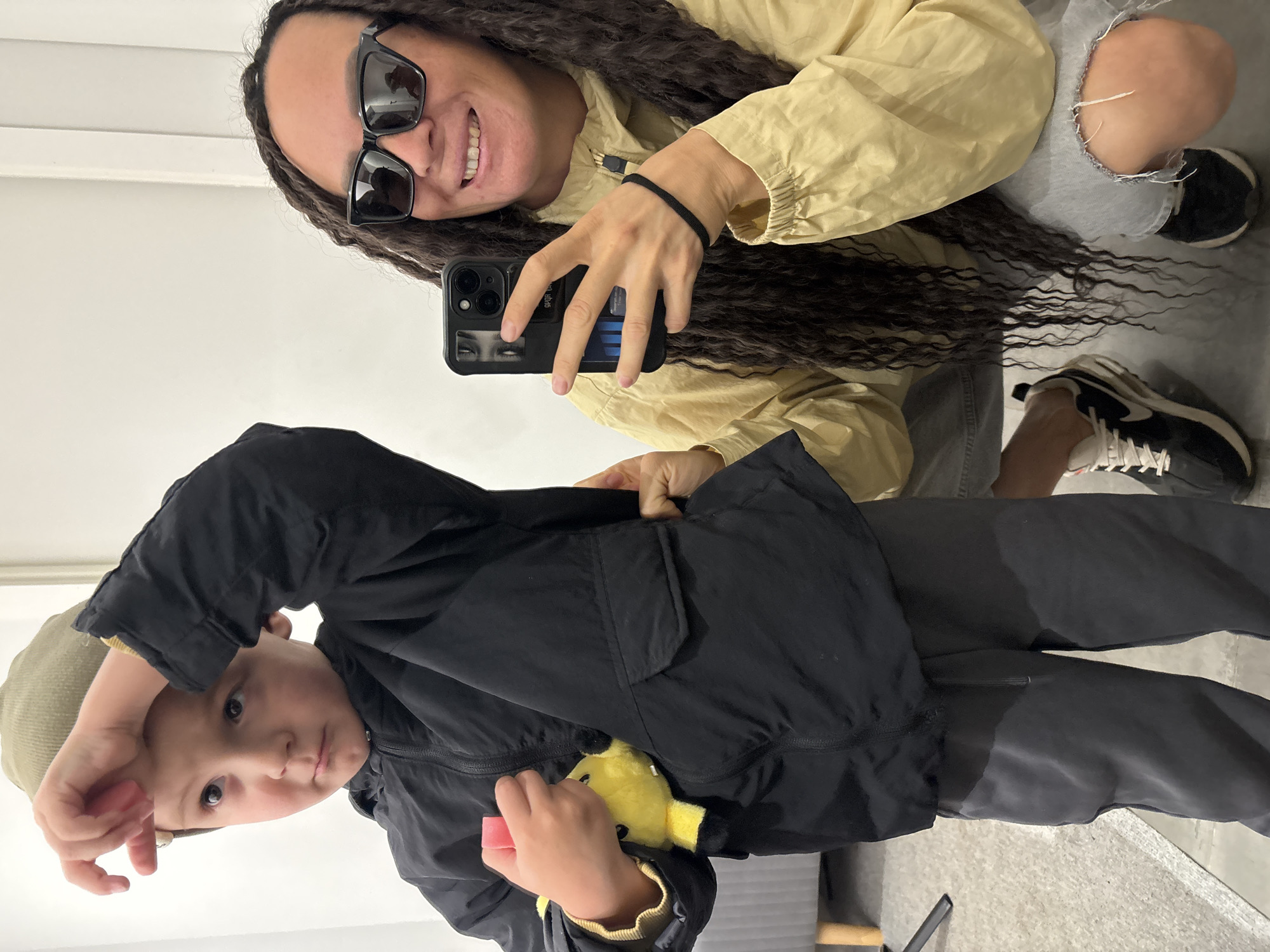 An indoor mirror selfie taken in Chaikovsky, Russia, features a woman and a child. The woman, positioned towards the right of the frame, is smiling and has long, dark, wavy hair. She wears black rectangular sunglasses, a light yellow long-sleeved jacket, and light-colored ripped jeans, with black and white sneakers visible on her feet. A black hair tie is on her left wrist. She holds a black smartphone with a distinctive case featuring eyes and blue elements in her right hand, taking the photograph.
To the left, a child, likely a boy, stands facing the camera with a neutral, slightly curious expression. He wears a black zipped jacket, dark pants, and a light olive-green ribbed beanie. He is holding a small yellow plush toy (resembling Pikachu) that peeks out from his left hand, which also holds a small pink object. The background is simple and bright, consisting of plain white walls and a light-colored floor, suggesting a casual setting, possibly a dressing room or home. The image captures a candid, everyday moment between the two subjects.