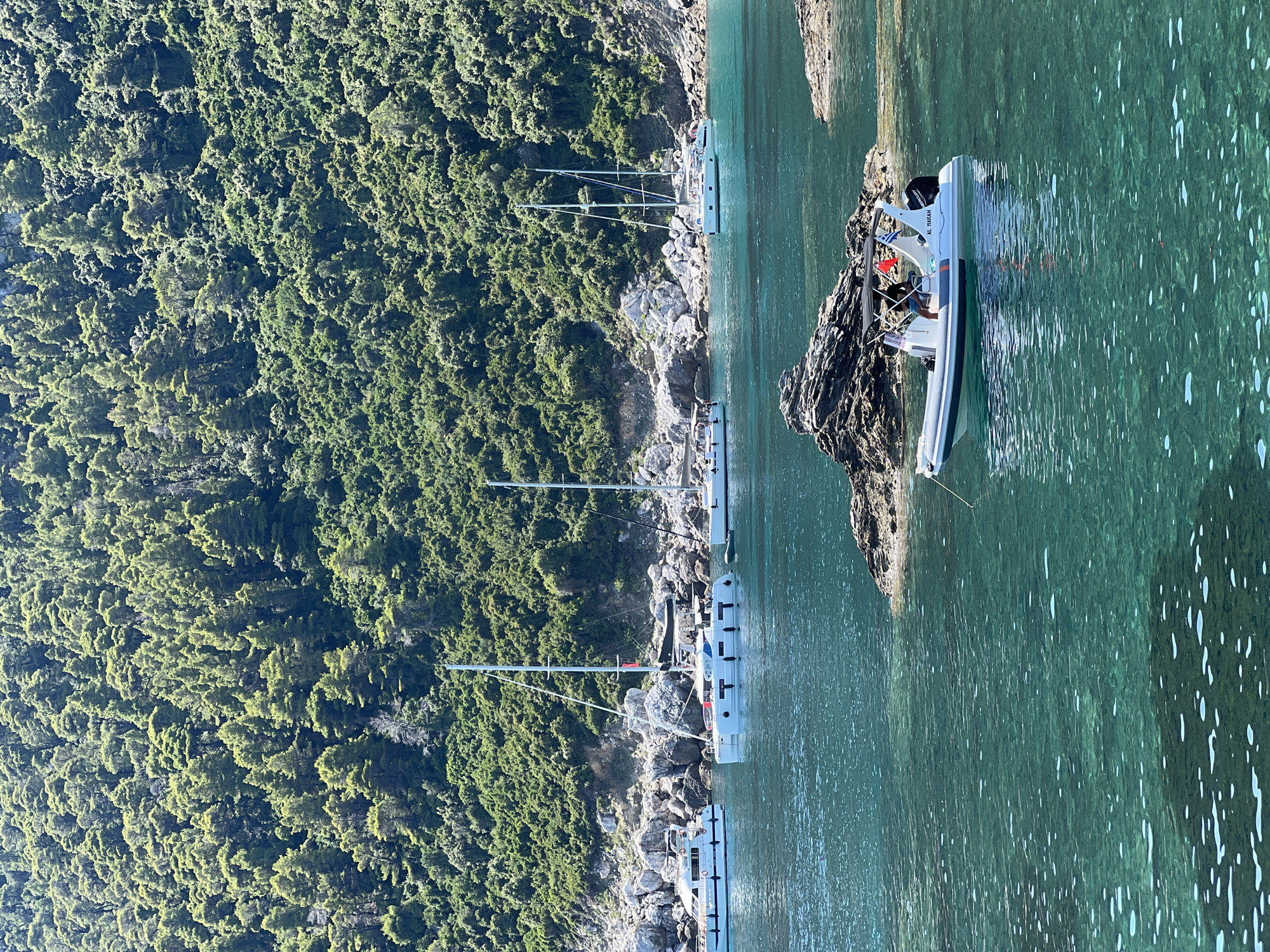 The image depicts a serene coastal scene in Loutraki, Greece. The foreground features crystal-clear turquoise water, with sunlight creating dappled patterns on the seabed. A white inflatable boat with a black canopy is anchored in the mid-ground, carrying two individuals, one of whom is steering. The boat is labeled "AL MARAH". In the background, several white sailboats are anchored, their masts reaching towards a lush, green hillside densely covered with trees. Rocky outcrops punctuate the shoreline. The overall impression is one of tranquility and natural beauty.