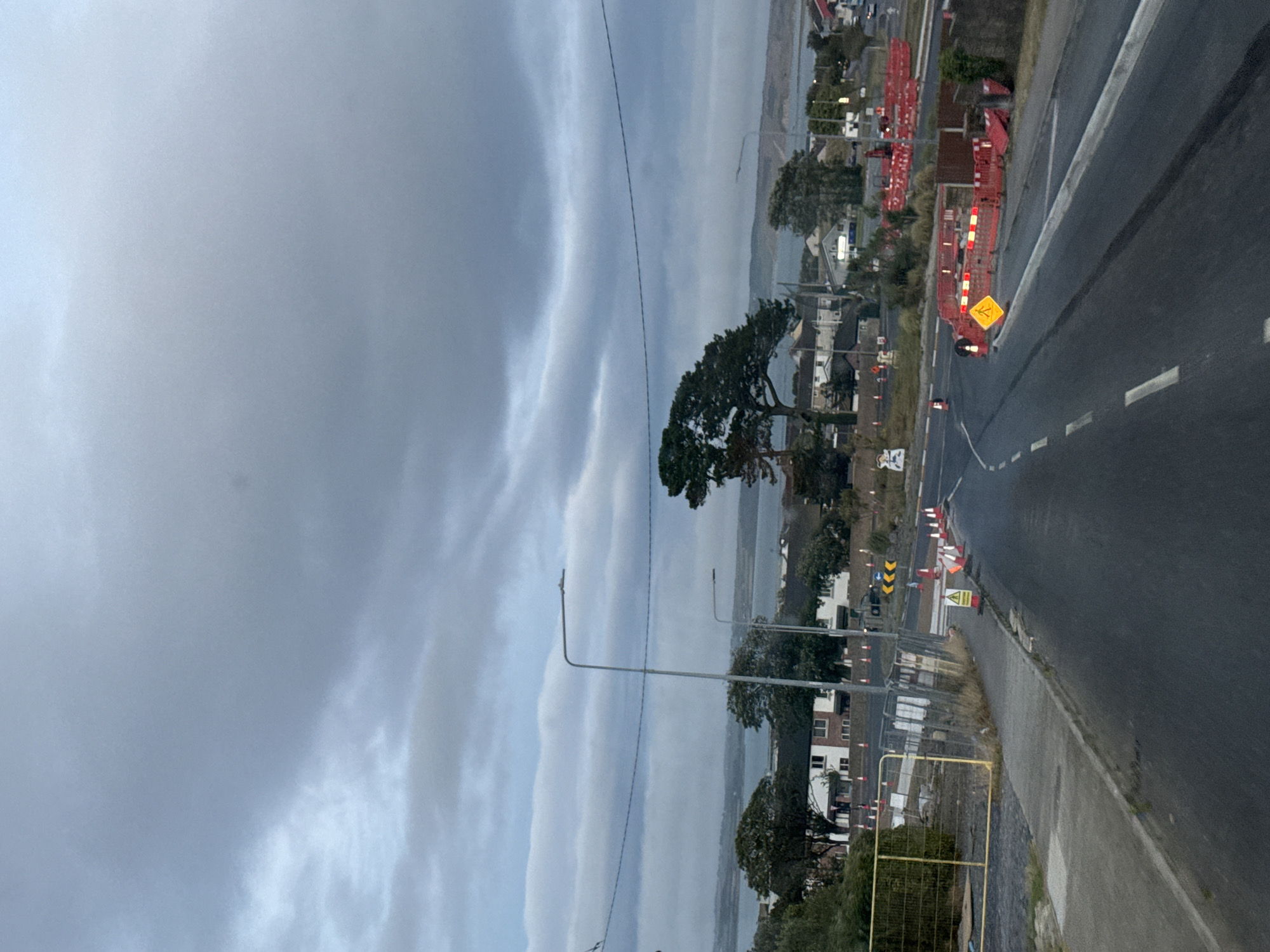 The image depicts a road in Tramore, Ireland, with ongoing construction work. The sky is overcast and gray. In the foreground, the asphalt road has white lane markings. To the left of the road, there is a sidewalk and a construction site enclosed by a yellow metal fence. Various orange traffic cones and red and white barrier fences are placed along the road, indicating a diversion or lane closure. A yellow diamond-shaped sign with a pedestrian symbol is visible near the barriers. In the background, there are buildings with white facades and dark roofs, some trees, and a body of water, likely Tramore Bay, with hills in the distance. There are no people visible in the image.