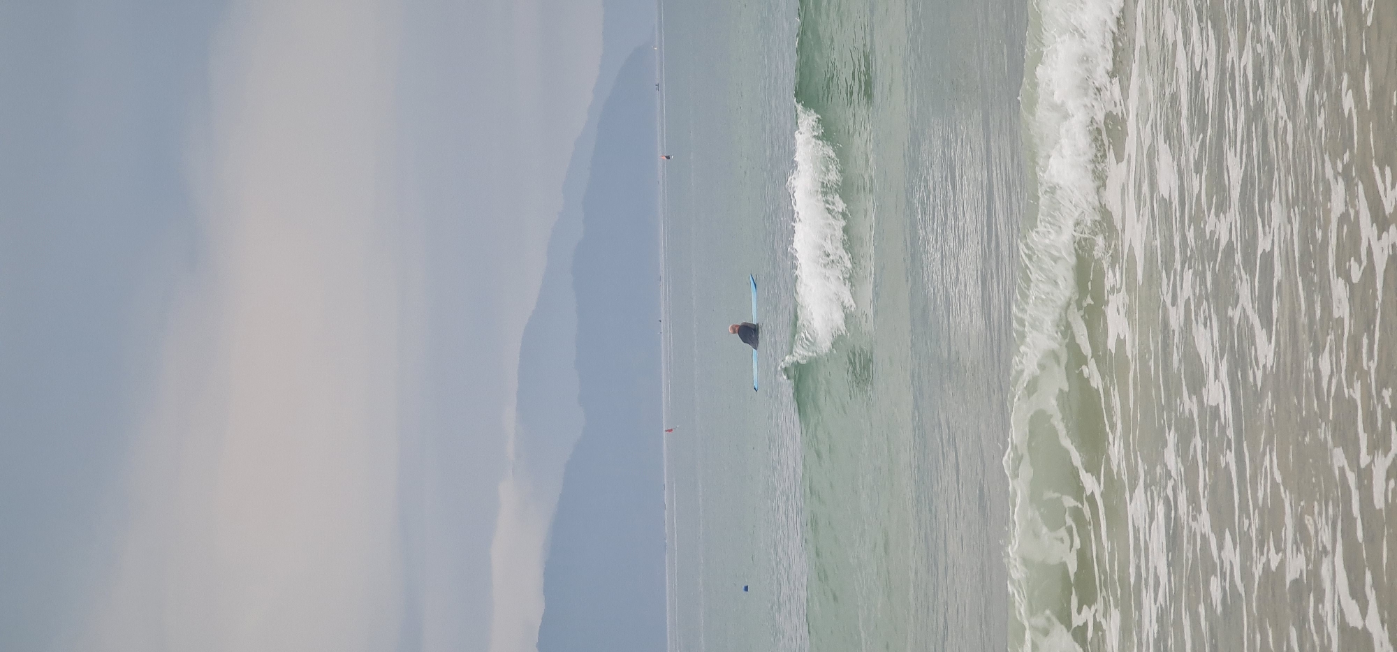 A photograph, taken at a distance and from a high vantage point, depicts a surfer in the ocean off the coast of Đà Nẵng, Vietnam. The surfer, positioned on a blue surfboard, is surrounded by a breaking wave. The water transitions from calmer, light-green hues to foamy white near the shore. In the background, a hazy, mountainous landmass is visible. Buoys are sparsely scattered across the water. The sky is overcast with a mix of grey and light-blue tones.
