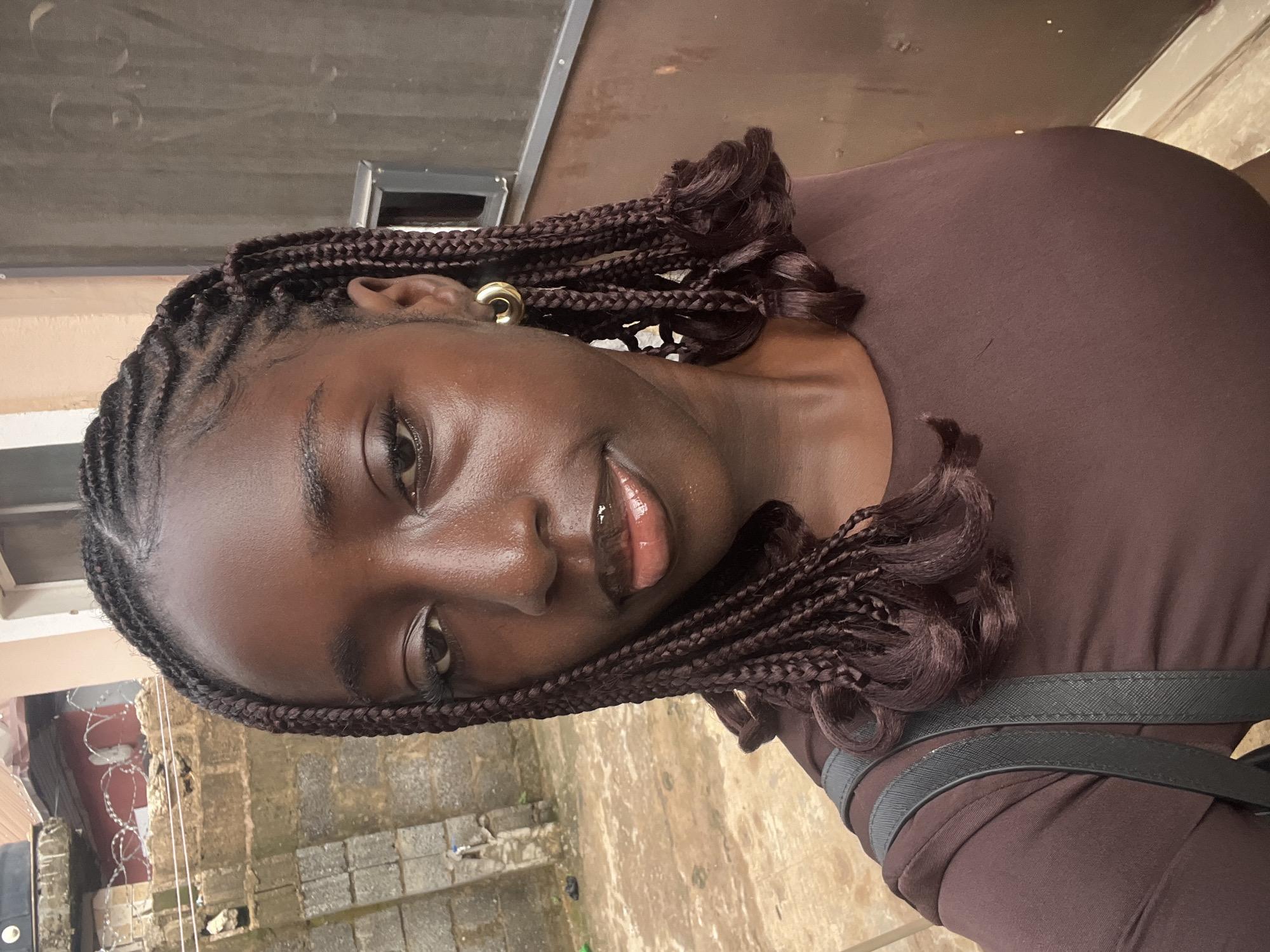 A dark-skinned woman with braided hair is looking at the camera with a slight smile. She is wearing a brown top. A black purse strap is visible across her shoulder. The background includes a wall and paved ground. It is difficult to definitively pinpoint a specific location in Abuja from this image alone.