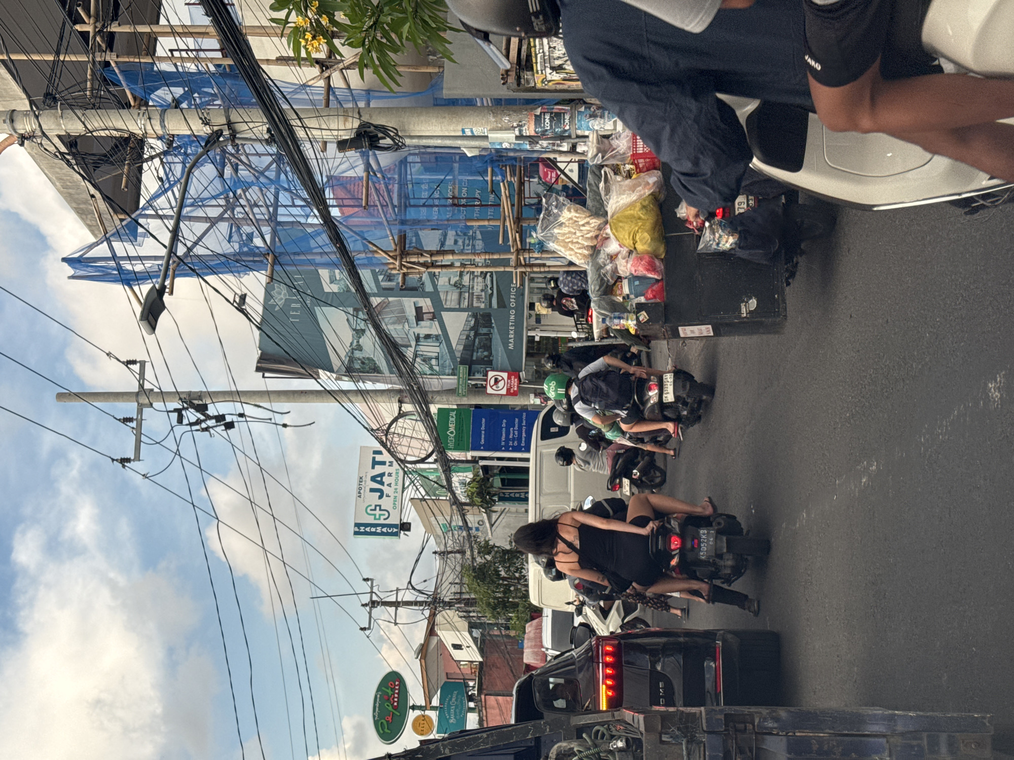 The scene is a busy street in what appears to be North Kuta, Indonesia. Traffic is congested with motorcycles and cars, with a woman on a motorcycle in the foreground. Street signage is visible for Jati Farm Pharmacy, Pepato Bakery, and Hydromedical Services. Overhead, there is a dense network of power lines. Scaffolding and construction materials are present, and blue netting is visible. There are advertising signs for real estate properties (Marketing Office). Pedestrians are visible amongst the traffic.