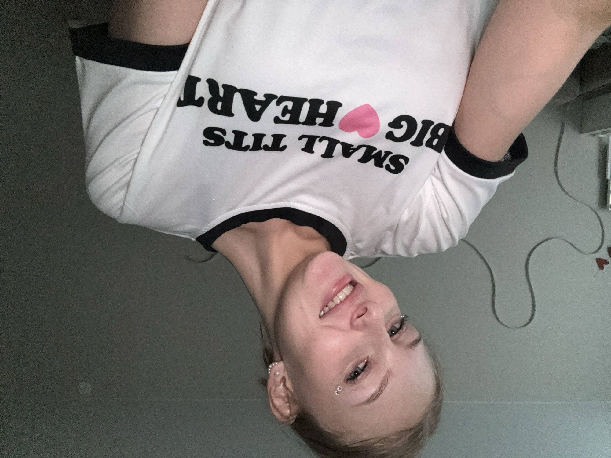 A young woman with blonde hair pulled back is smiling at the camera. She is wearing a white t-shirt with black trim around the collar and sleeves. The t-shirt has black text on it that reads "SMALL TITS BIG HEART" with a pink heart in place of the word "BIG". She has several small silver piercings on her face, including one above her eyebrow and others on her cheekbone. She also has earrings. The background is a lightly colored wall with some faint, dark grey wavy lines and a few small red heart shapes painted on it. The lighting is dim. The image appears to be a selfie taken indoors, possibly in a bedroom. There is no indication of the location being the city of Perm, Russia.