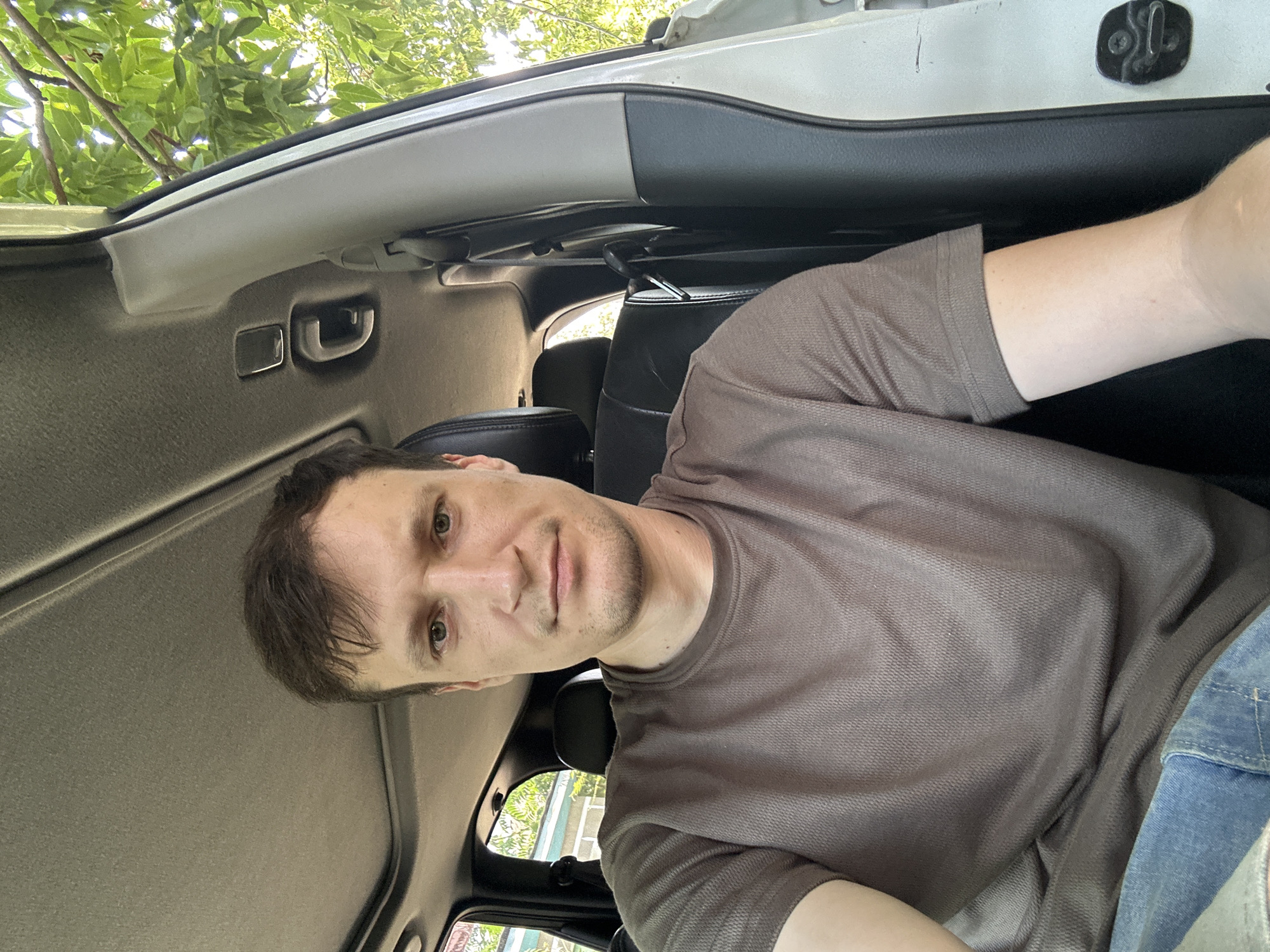 A medium shot photo shows a fair-skinned man with short, dark brown hair and light green eyes sitting in the driver's seat of a vehicle. He is wearing a brown t-shirt and blue jeans. The man is looking directly at the camera with a neutral expression. The interior of the vehicle is visible, including the car's roof and ceiling with a sunroof. Outside the car, through the open passenger door and window, green foliage and trees are visible, suggesting an outdoor setting. The background on the left side of the image shows a portion of a building with a light-colored wall and a window, suggesting the vehicle may be parked near a residential area. The location is identified as Makiv, Ukraine.