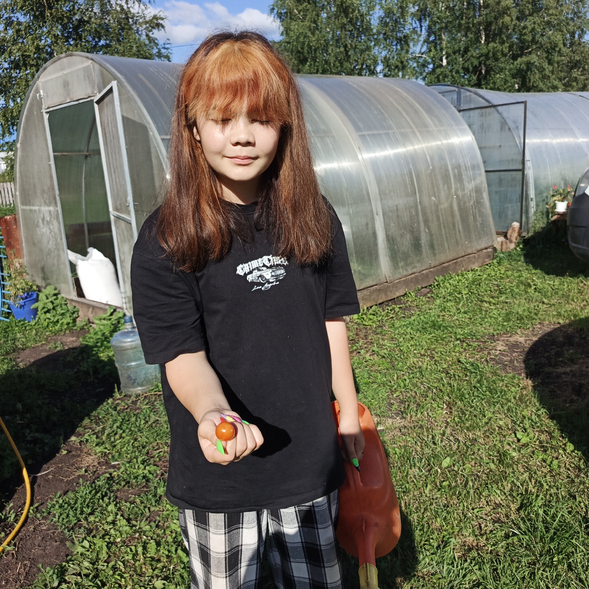 In Podlubovo, Russia, a young person with auburn and blonde-streaked hair, eyes closed, stands in a grassy yard. They are wearing a black t-shirt with a graphic of a car and text, and black and white checkered pants. The person holds a small, round, orange object, possibly a tomato, in their right hand, which has brightly colored nail polish. A large orange watering can is held in their left hand. Behind the person are two clear plastic greenhouses, with one having an open door revealing a large white bag inside. Trees and a bright blue sky are visible in the background. The scene appears to be set on a sunny day.