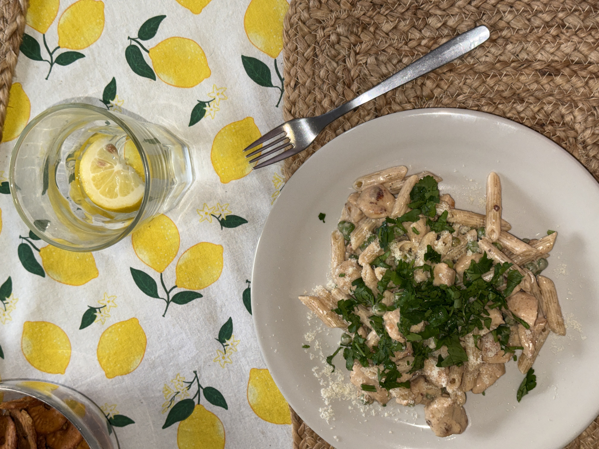 The image displays a top-down view of a dining table set for a meal. A white tablecloth, adorned with a repeating pattern of bright yellow lemons, green leaves, and small white flowers, covers the table.
On the right side of the frame, a round, light brown woven placemat, possibly made of jute, is visible. It holds a white, round ceramic plate featuring a generous serving of penne pasta. The pasta is coated in a creamy, light-colored sauce and contains chunks of what appears to be chicken and green peas. The dish is heavily garnished with freshly chopped green herbs, likely parsley, and a sprinkling of grated white cheese. A silver fork rests diagonally on the woven placemat, positioned above the plate.
To the left, a clear glass of water sits directly on the lemon-patterned tablecloth. It contains a slice of lemon and a few ice cubes. In the bottom-left corner of the image, the edge of a glass bowl or container is visible, holding numerous golden-brown items, which could be crackers, bread chips, or a similar snack.
The scene suggests an inviting, casual indoor meal setting. There are no people visible, and no discernible text appears in the image. The lighting is consistent with an indoor environment.