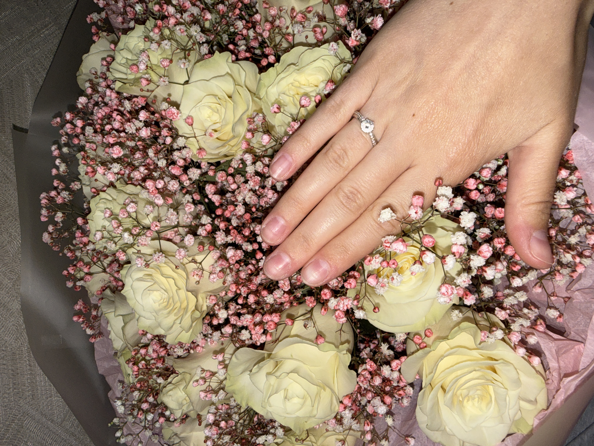 A hand adorned with a ring is placed over a bouquet of pale yellow roses and pink and white baby's breath. The background is a grey textured surface. The image does not contain any information related to the city of Chaikovsky, Russia.