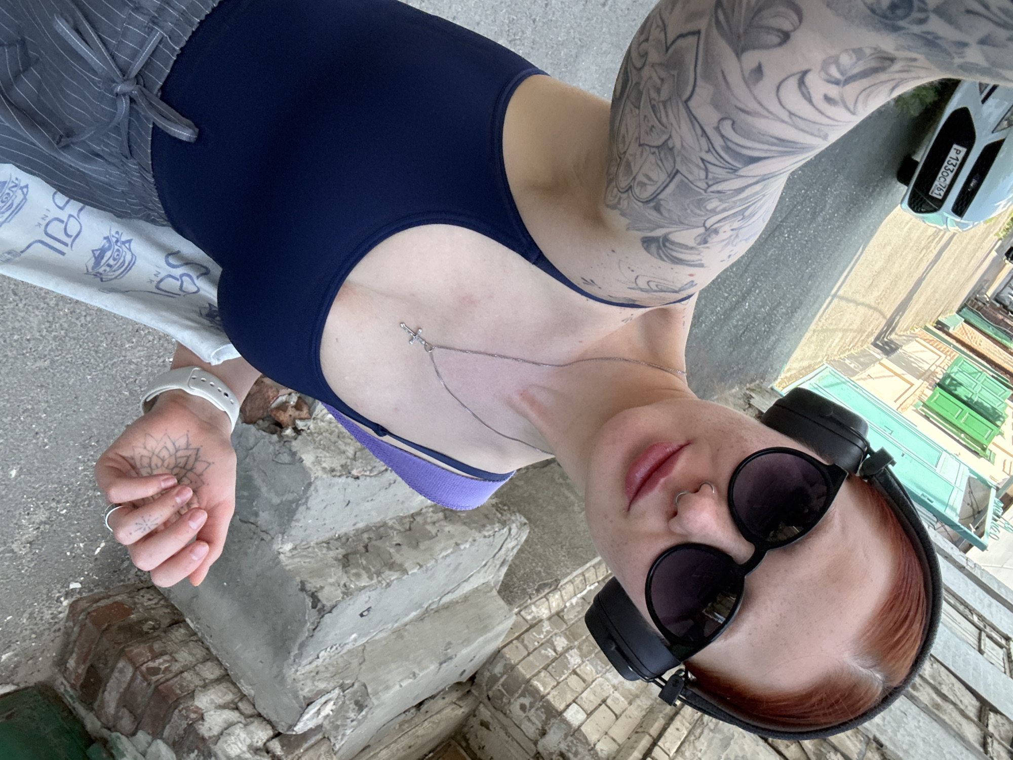 A woman poses for a selfie on a street in Rostov-on-Don, Russia. She wears a dark blue swimsuit-style top, striped gray pants, sunglasses, and headphones. She has tattoos on her arms and hands, and a silver necklace. The background shows a street with buildings, greenery, and a parked white car (plate number visible: P133OC/161). She is standing next to some crumbling steps.