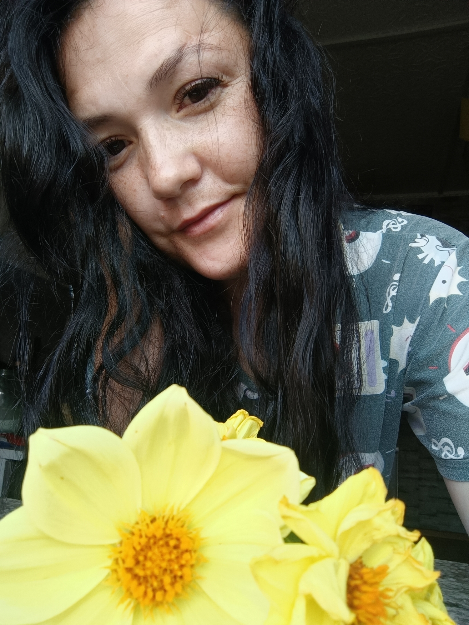 A woman with long, dark hair and freckled skin is shown in a close-up shot, holding yellow flowers in the foreground. She is wearing a dark green shirt with a white cartoonish print. Her hair is partially obscuring her face, and she is looking towards the camera with a slight smile. The background is dark and indistinct, suggesting an indoor setting. The image does not contain any discernible features that would indicate it was taken in Karsha, Russia.