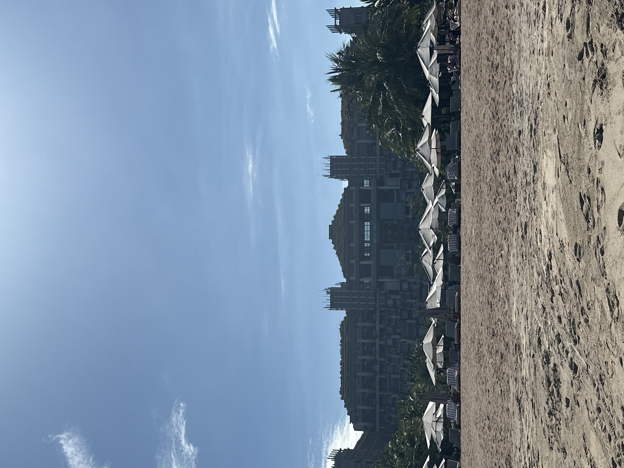 This image shows a beach scene in Kuta Selatan, Indonesia. The beach is sandy and the sky is blue with some clouds. A large building is visible behind the beach umbrellas and palm trees. The building appears to be a resort or hotel. Beach umbrellas and lounge chairs are positioned on the sand. The image captures a tourist destination.