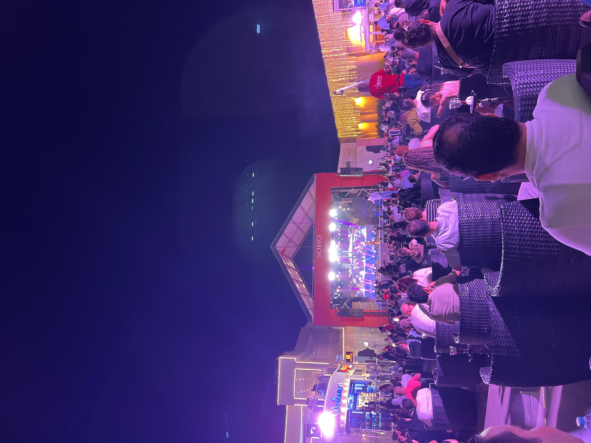 The image depicts an outdoor evening event at Soho Square in Sharm El Sheikh, Egypt. A stage with a large screen is set up, featuring a live performance with musicians and a singer, illuminated by stage lights. A crowd of people is seated in an open area facing the stage, some at tables with what appear to be shisha pipes. Surrounding the square are illuminated buildings with architectural details, and strings of golden lights hang from one of the structures. The sky above is a deep, dark blue, indicative of nighttime. People are dressed casually, and the atmosphere appears to be festive. A man in a red shirt with "EGYPT ENTERTAINMENT" on the back is visible near the stage area.