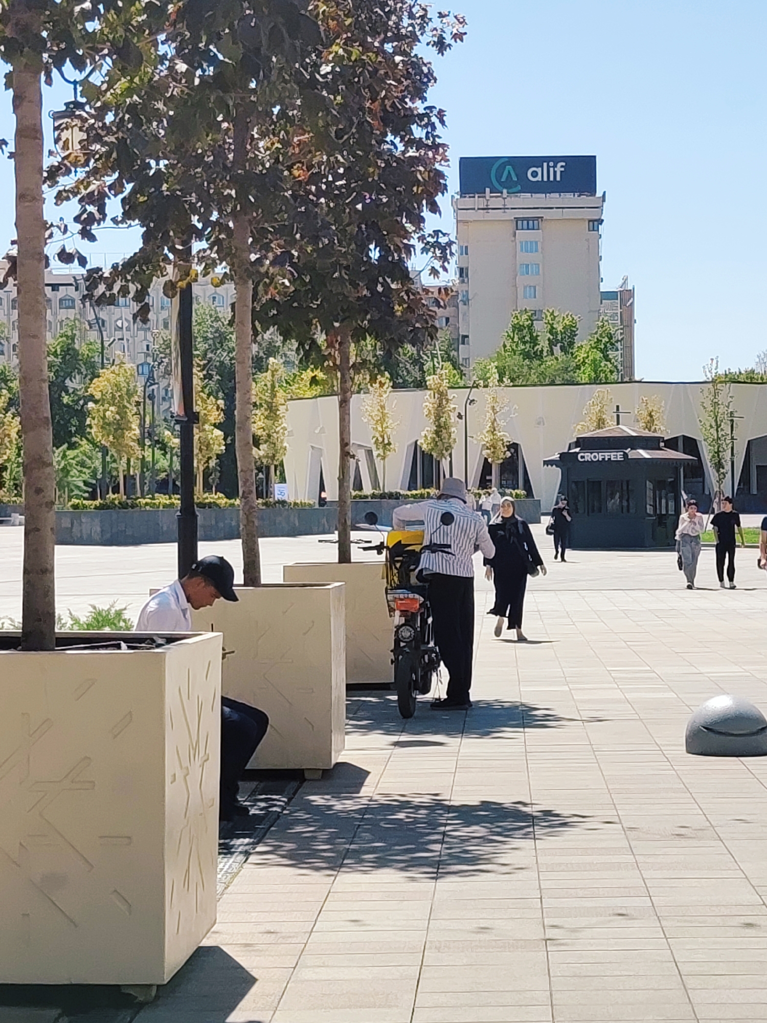 The image depicts an outdoor scene in Tashkent, Uzbekistan. In the foreground, a man wearing a white shirt and a black baseball cap is seated next to a large, decorative planter. His body is partially obscured by the planter, and he appears to be looking down. To his right, another man wearing a hat, a striped shirt, and dark pants is standing beside a parked scooter with a yellow cargo box.
In the mid-ground, several people are walking across a paved plaza. One woman is wearing a black headscarf and a dark abaya. Further back, a small kiosk with the word "CROFFEE" visible on its signage is situated in front of a modern building with white, angular architectural features.
In the background, a tall building is topped with a prominent sign displaying the logo and name "alif." The sky is clear and blue. The overall environment suggests a public urban space, likely a park or pedestrian area, during daylight.
