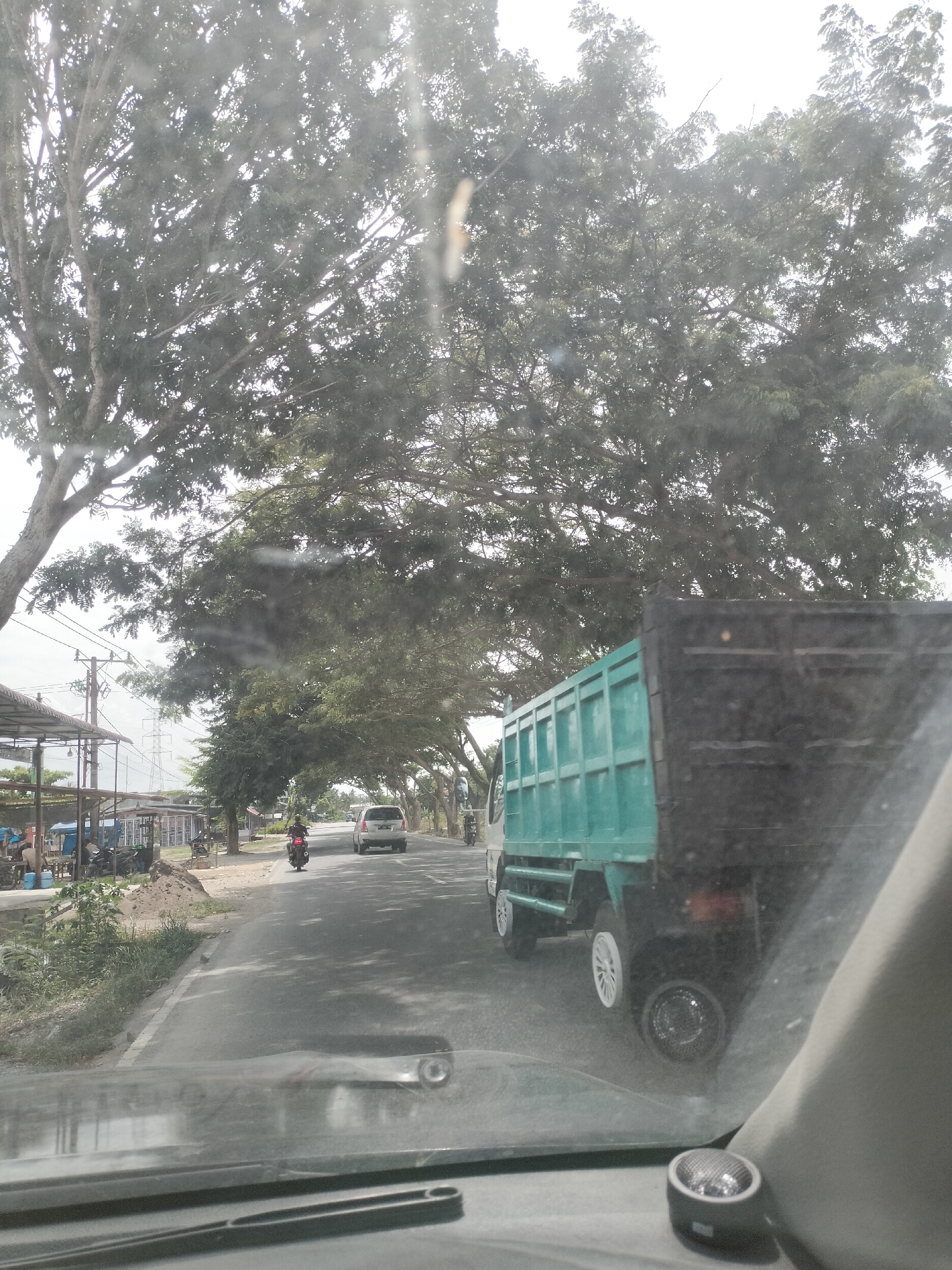 A truck with a turquoise bed and black cab is on the right side of the road, partially obscuring the view. In the distance, a white car and a motorcycle are also on the road. On the left side of the road, there are buildings with awnings and parked motorcycles. Tall trees with lush green foliage line both sides of the road, creating a canopy overhead. The sky is bright and overcast, suggesting a daytime scene in Putoh Sa, Indonesia. The image appears to be taken from inside a vehicle, looking out through the windshield, which has some smudges and reflections.