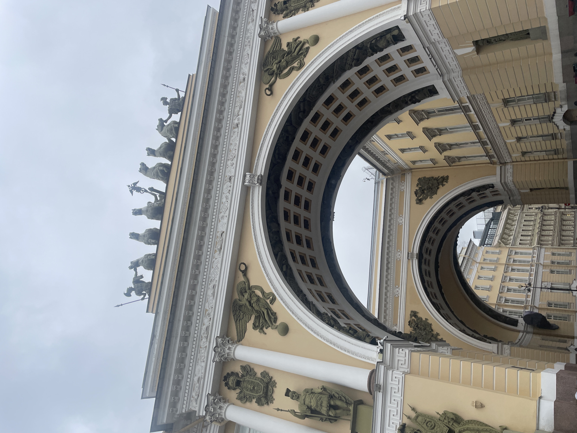 The image depicts the General Staff Building arch in Palace Square, Saint Petersburg, Russia, under a cloudy sky. The grand yellow archway features intricate architectural details, including columns, relief sculptures of winged figures, and a chariot pulled by horses crowning the top. The arch's interior is adorned with square panels. Beyond the arch, multiple stories of yellow buildings with numerous windows are visible. The scene is devoid of any people, emphasizing the monumental architecture. The lighting is diffused due to the overcast conditions.