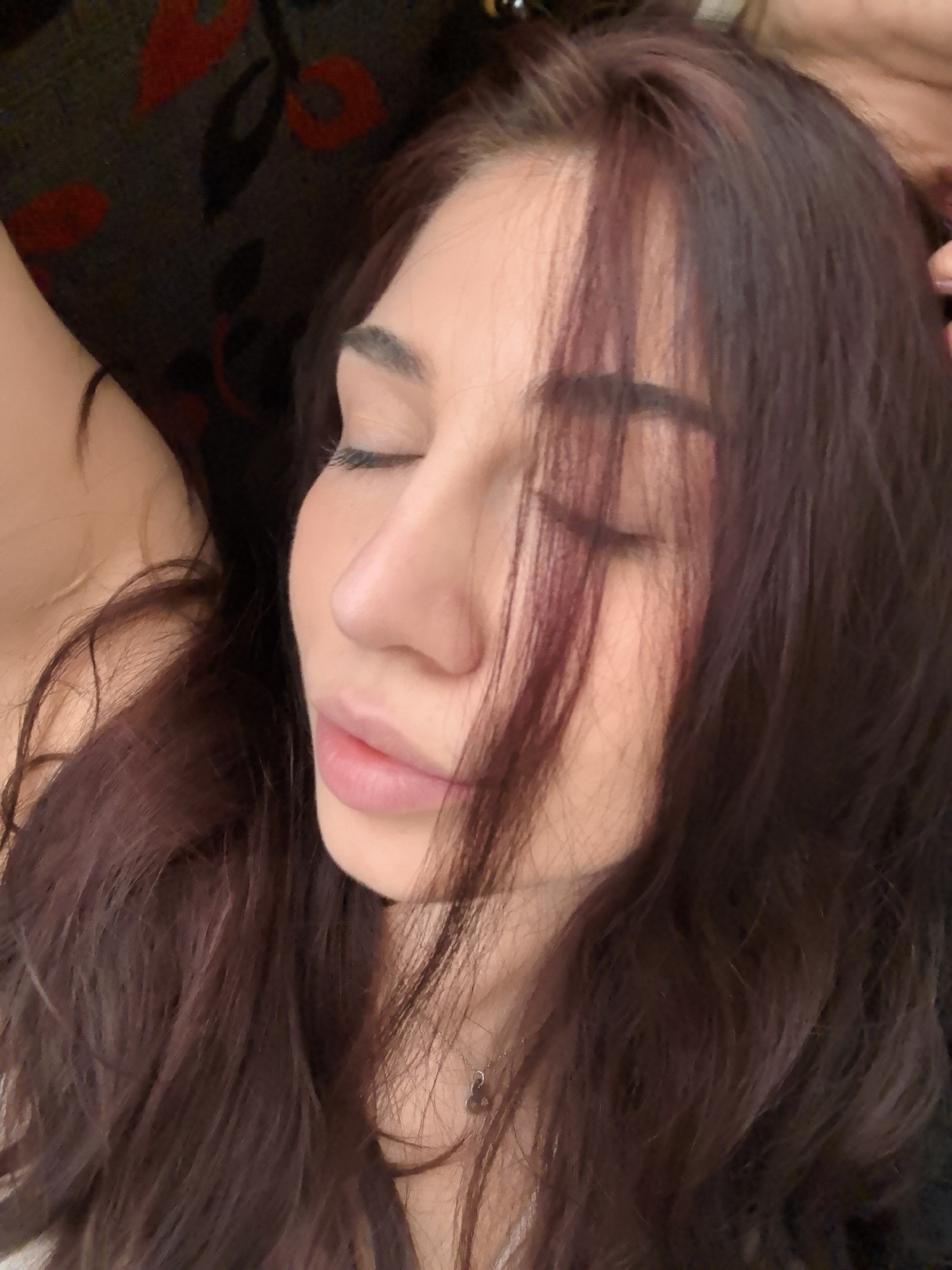 A close-up shot captures a woman with dark auburn hair lying down with her eyes closed. Her complexion appears fair. A sliver of her shoulder is visible on the left side. The background behind her suggests a dark surface with patterns, possibly a couch or seat. The details of a specific location in Stara Zagora, Bulgaria, are not discernible from the image.