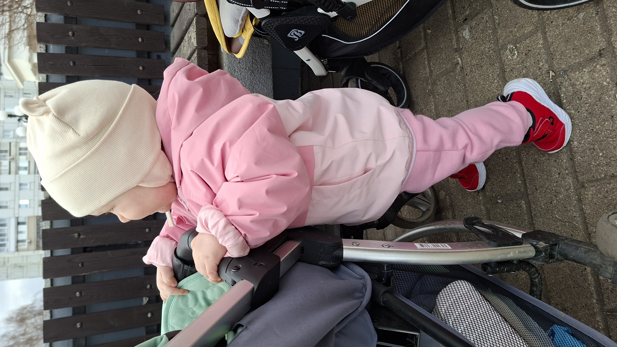 A toddler in Chaikovsky, Russia, wearing a cream-colored knit hat, pink two-tone snowsuit, and red and white sneakers, stands next to a stroller on a paved surface. The stroller, containing a gray storage basket and green lining, is positioned near a dark brown slatted bench in front of a multi-story building. A second stroller is partially visible in the upper right corner of the image.