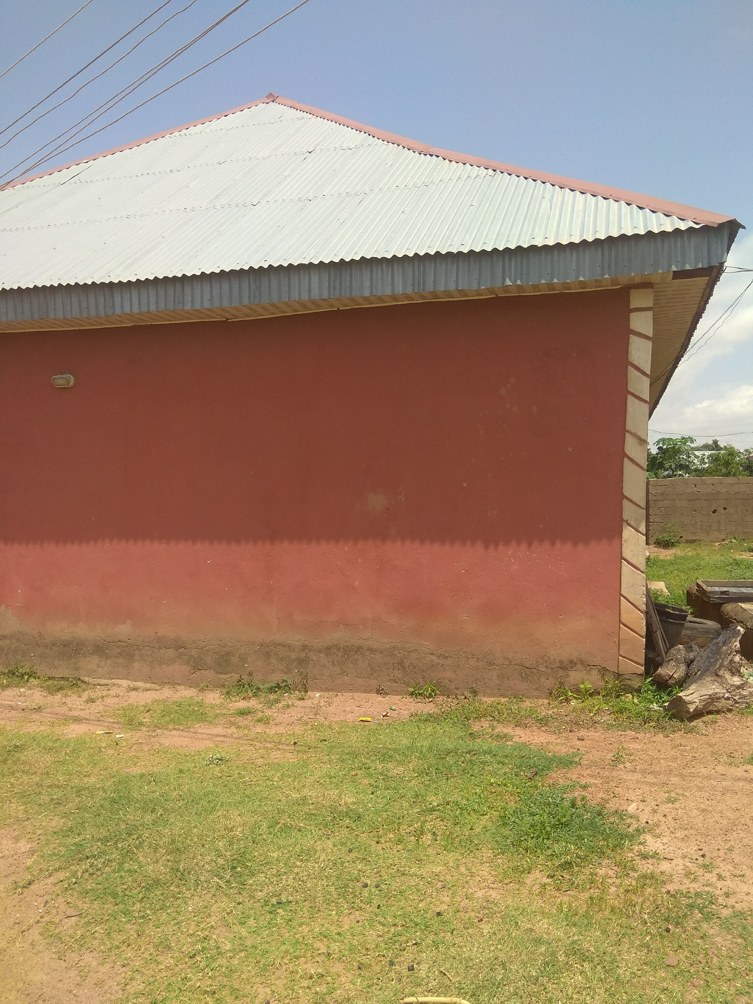 The image depicts the side view of a red-walled building with a corrugated metal roof in Jalingo, Nigeria. The building has a light fixture on its upper left wall, and the base of the wall is weathered and stained. A strip of decorative stonework runs vertically along the right edge of the wall. The ground in front of the building is a mix of dry earth and sparse green grass. In the background, beyond a low stone wall, trees and the sky are visible. To the right of the building's corner, there are some discarded items including logs and tires. The overall scene suggests an outdoor environment in a semi-urban or rural setting.