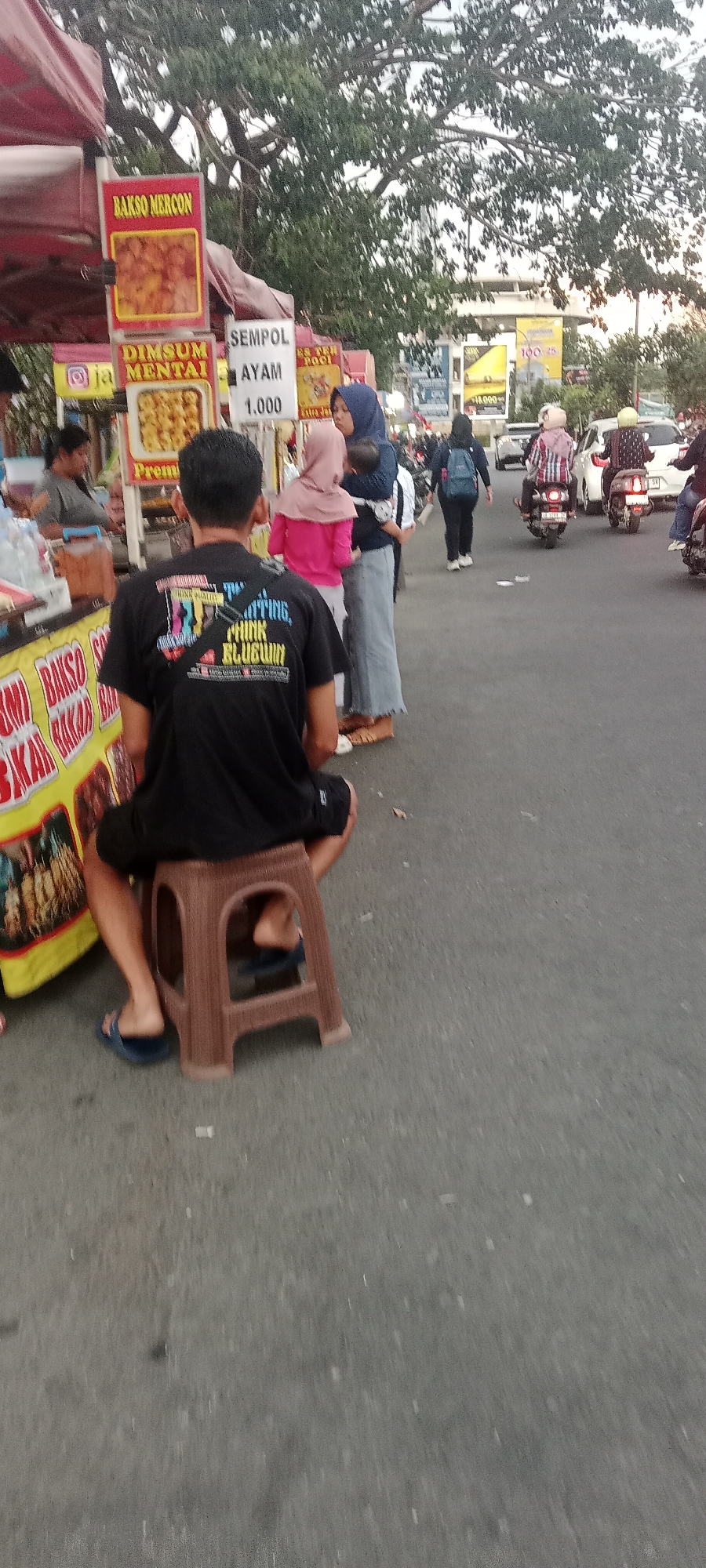 The image depicts a street scene in Ngemplak, Indonesia, at dusk. The foreground is dominated by a person wearing a black t-shirt and shorts, sitting on a brown plastic stool. Their back is to the camera, and they are wearing blue flip-flops. To the left, a yellow food stall displays various food items, including "Bakso Mercon" and "Dimsum Mentai". Behind the person, several other food stalls are visible, with signs indicating "Sempol Ayam" and prices like "1.000". Several people are walking on the street, including women wearing hijabs and carrying children, and others on motorcycles. The street appears to be busy with pedestrian and vehicle traffic. In the background, there are buildings and trees, with the sky suggesting the late afternoon or early evening. The overall impression is a lively street food scene in a populated urban or semi-urban area.