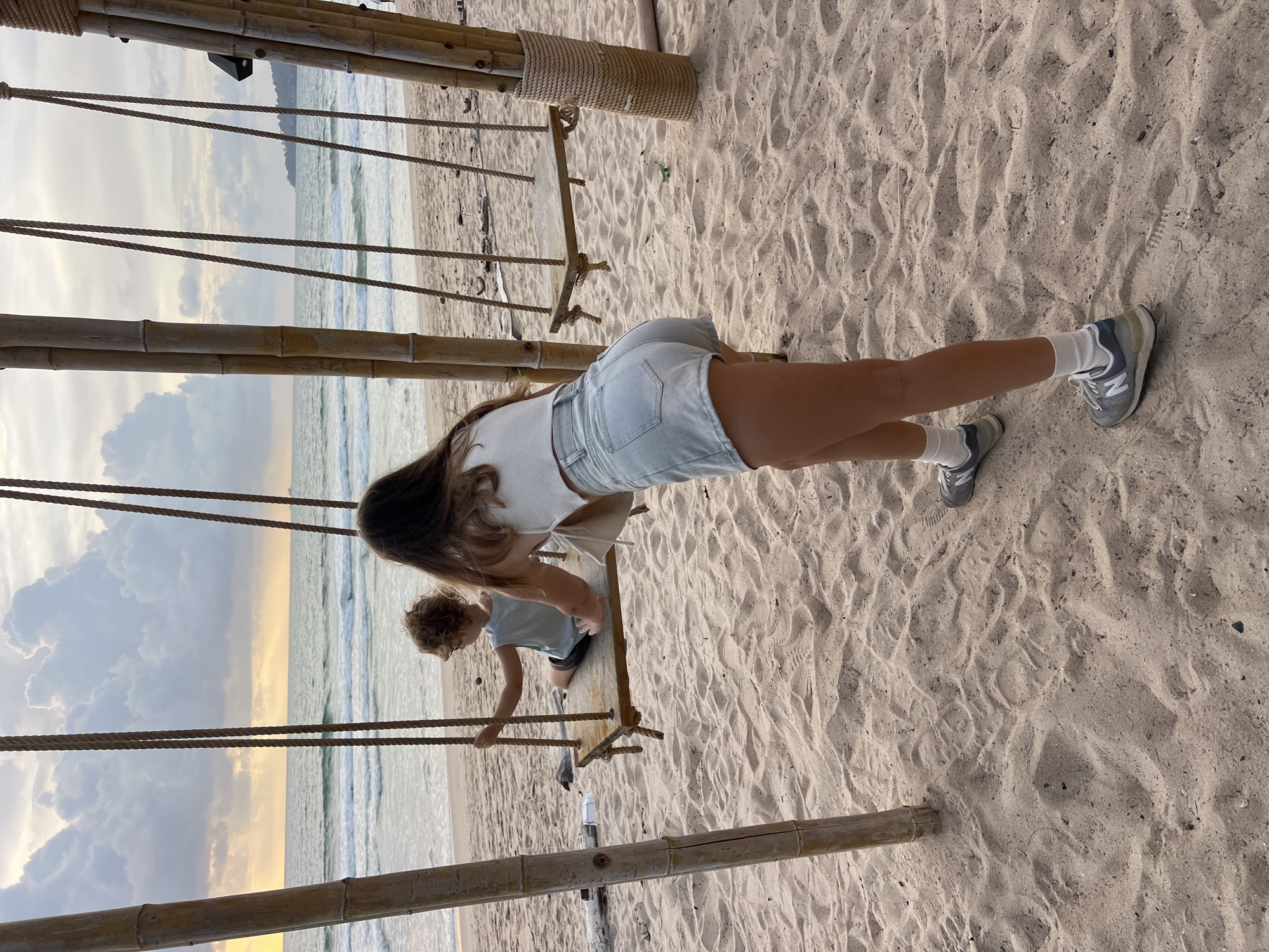 A young child sits on a rustic swing made of wood and rope, suspended from a bamboo frame on a sandy beach in Choeng Thale, Thailand. The child is facing away from the camera, looking out at the ocean as gentle waves lap the shore under a cloudy sky with hints of sunset. Behind the child, a woman with long brown hair, wearing a white crop top and light denim shorts, leans forward, her right hand extended towards the swing, suggesting she might be pushing it. She is dressed casually in gray athletic shoes with white socks. The beach is a mix of fine sand with some debris, and the overall atmosphere is tranquil, with the ocean extending to the horizon.