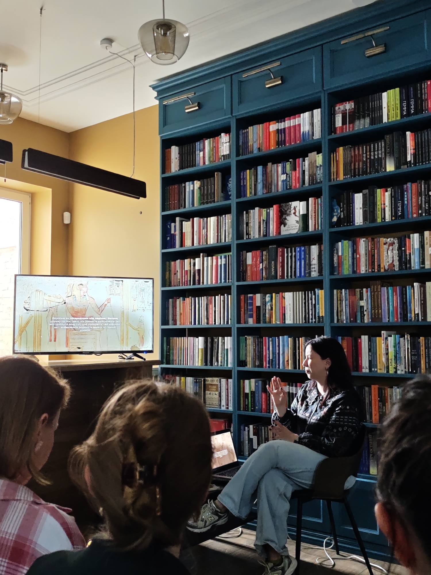 The photograph captures a scene in what appears to be a library or study, likely in Vinnytsia, Ukraine. The setting is characterized by a large, blue built-in bookcase filled with books, which serves as a prominent backdrop to the scene. In the foreground, an unseen group of people is listening attentively to a woman who is seated on a chair and appears to be giving a presentation or lecture. She is dressed in a patterned shirt and jeans, with her legs crossed and one hand raised in a gesture, suggesting she is actively speaking. A laptop rests on a chair near her.
In the background, a television screen displays an image depicting ancient Egyptian figures and hieroglyphs, indicating that the presentation likely involves the topic of ancient Egypt. Above the television, there is a long, rectangular light fixture. The room is illuminated by natural light coming from a window on the left and supplemented by overhead lighting fixtures, including suspended pendant lamps. The walls are painted in warm, earthy tones. The overall atmosphere suggests an educational or cultural event taking place in a library setting.