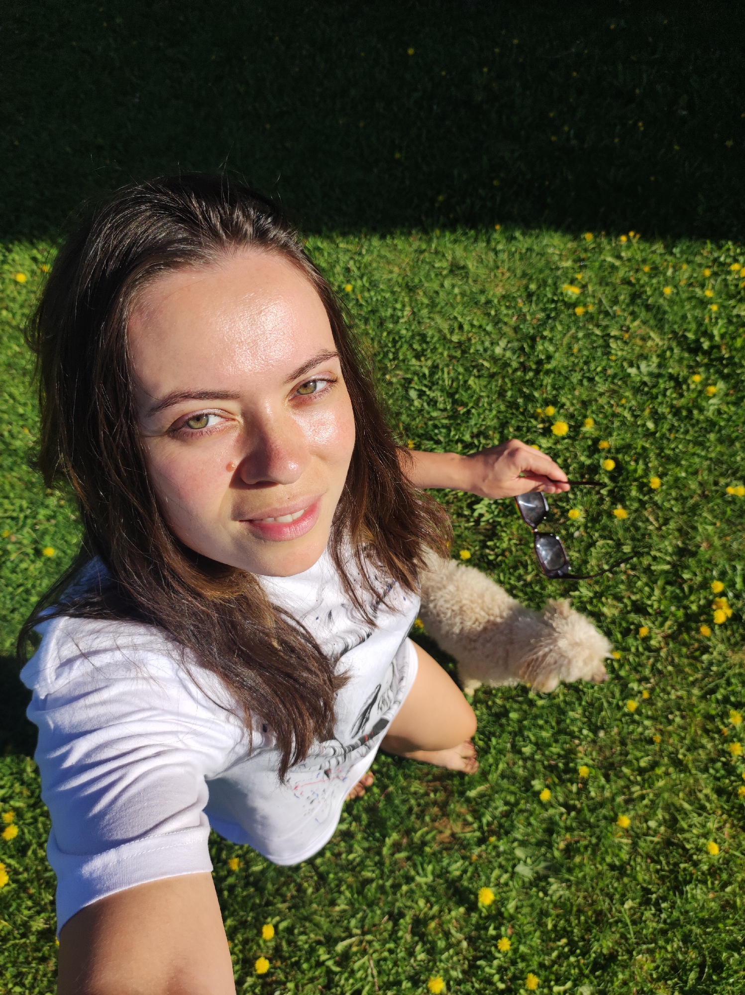 A high-angle, daytime shot shows a woman and her dog outdoors in a grassy area. The woman, who is fair-skinned with brown hair and green eyes, is looking directly into the camera. She wears a white t-shirt. Her right hand is holding a pair of dark sunglasses. Beside her stands a small, light-colored, possibly poodle-mix dog. The background consists of green grass dotted with yellow dandelions. The scene is well-lit, suggesting sunny weather. The location is unknown, however the city of Фоки, Russia was specified.