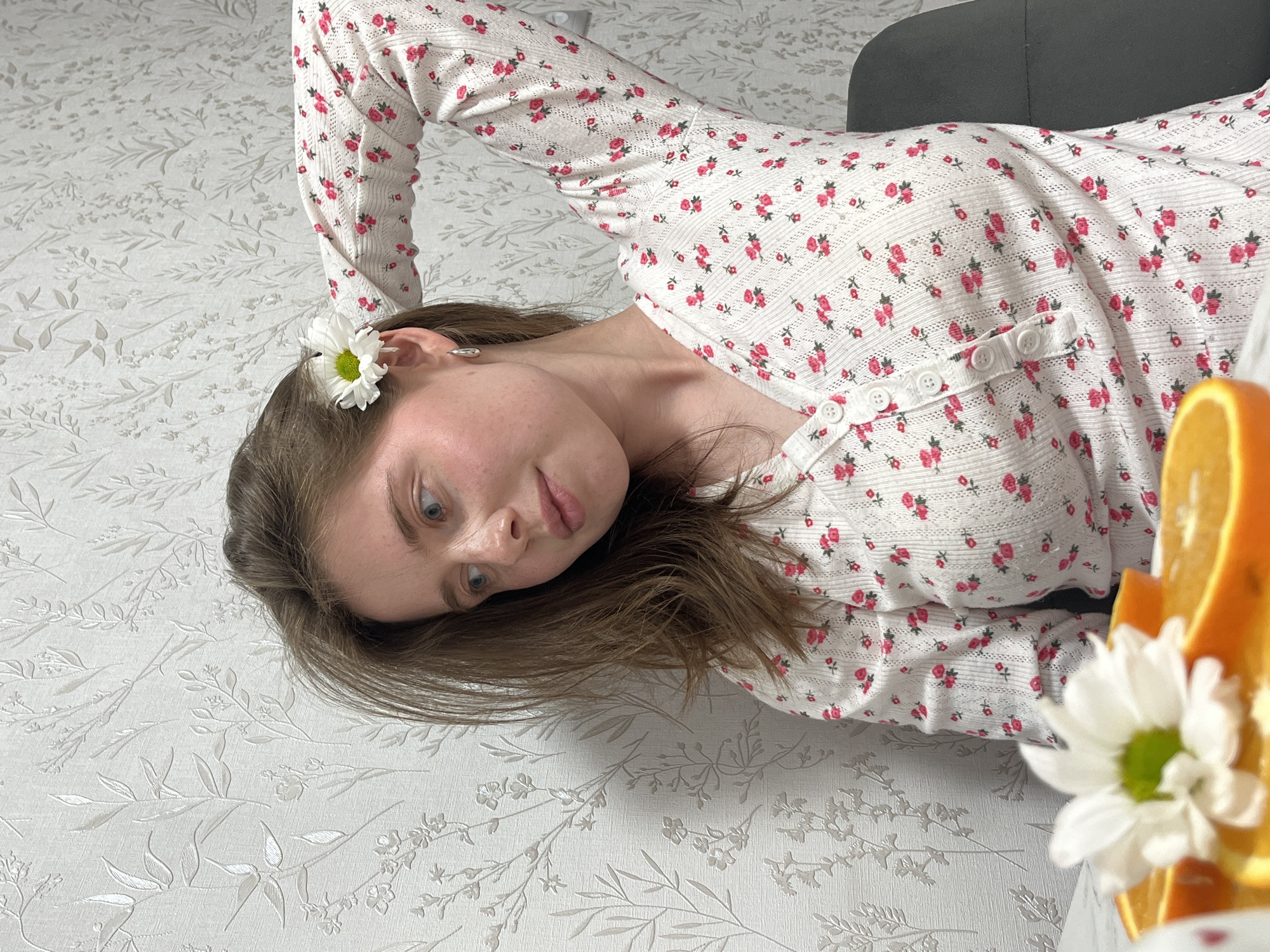 A woman with brown hair is lying on a white surface with a floral pattern. She is wearing a white, long-sleeved shirt with small pink floral designs and white buttons. A white daisy is tucked behind her left ear. An orange and another white daisy are visible in the foreground, partially obscuring the view. A grey object is in the upper right corner of the frame. The location is likely Chaikovsky, Russia, though the context of this setting within the image is not clearly defined.