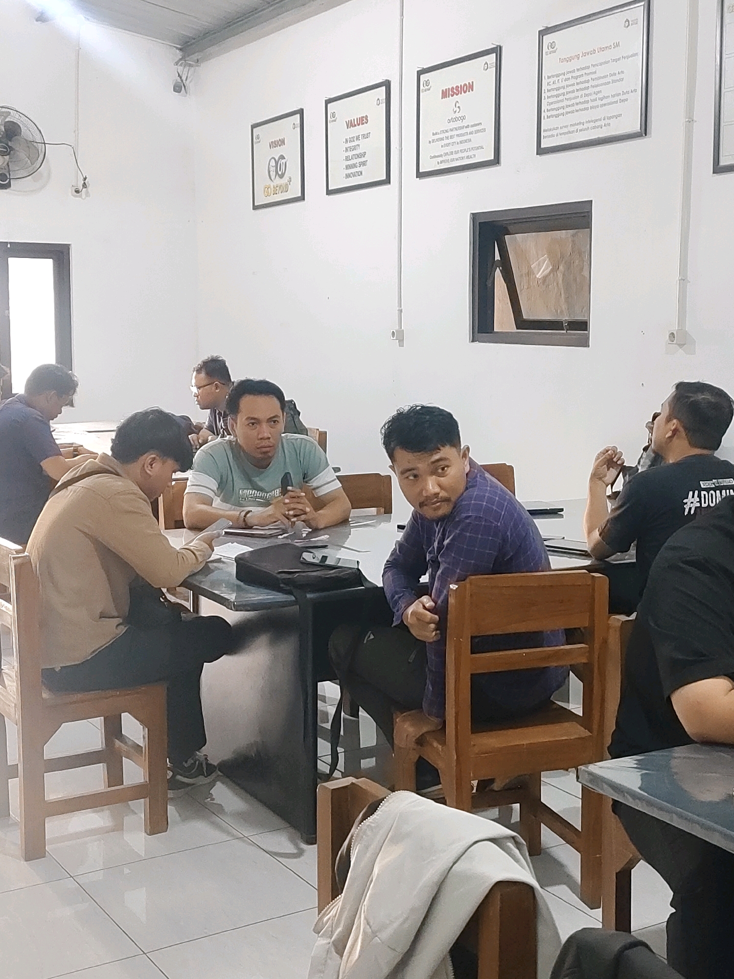 This image depicts an indoor gathering, possibly a meeting or a casual get-together, in a room with white walls and tiled flooring. In the background, there are framed posters on the wall displaying text and graphics, some of which appear to be company values or mission statements. A fan is visible on the left side of the frame.
In the foreground, several individuals are seated around a dark-colored table. The lighting suggests an ambient indoor setting.
The people are dressed in casual attire. Some individuals are looking in different directions, while others appear to be engaged in conversation or observing something. There are also personal belongings such as bags and papers on the table.
Based on the context provided about the location being Probolinggo, Indonesia, it is plausible that this scene is within a business or community space in that city. The overall atmosphere seems relaxed yet focused, typical of informal meetings or brainstorming sessions.
