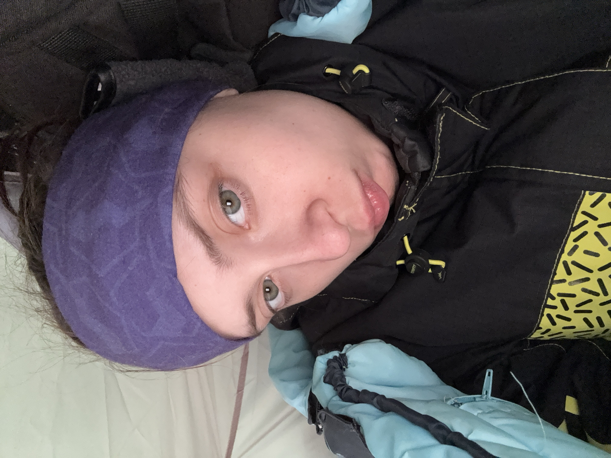 A person is lying in a tent or sleeping bag. The person has a purple headband and fair skin. They are wearing a black jacket with yellow accents. The sleeping bag or tent is light blue. The location is not discernible from the image.