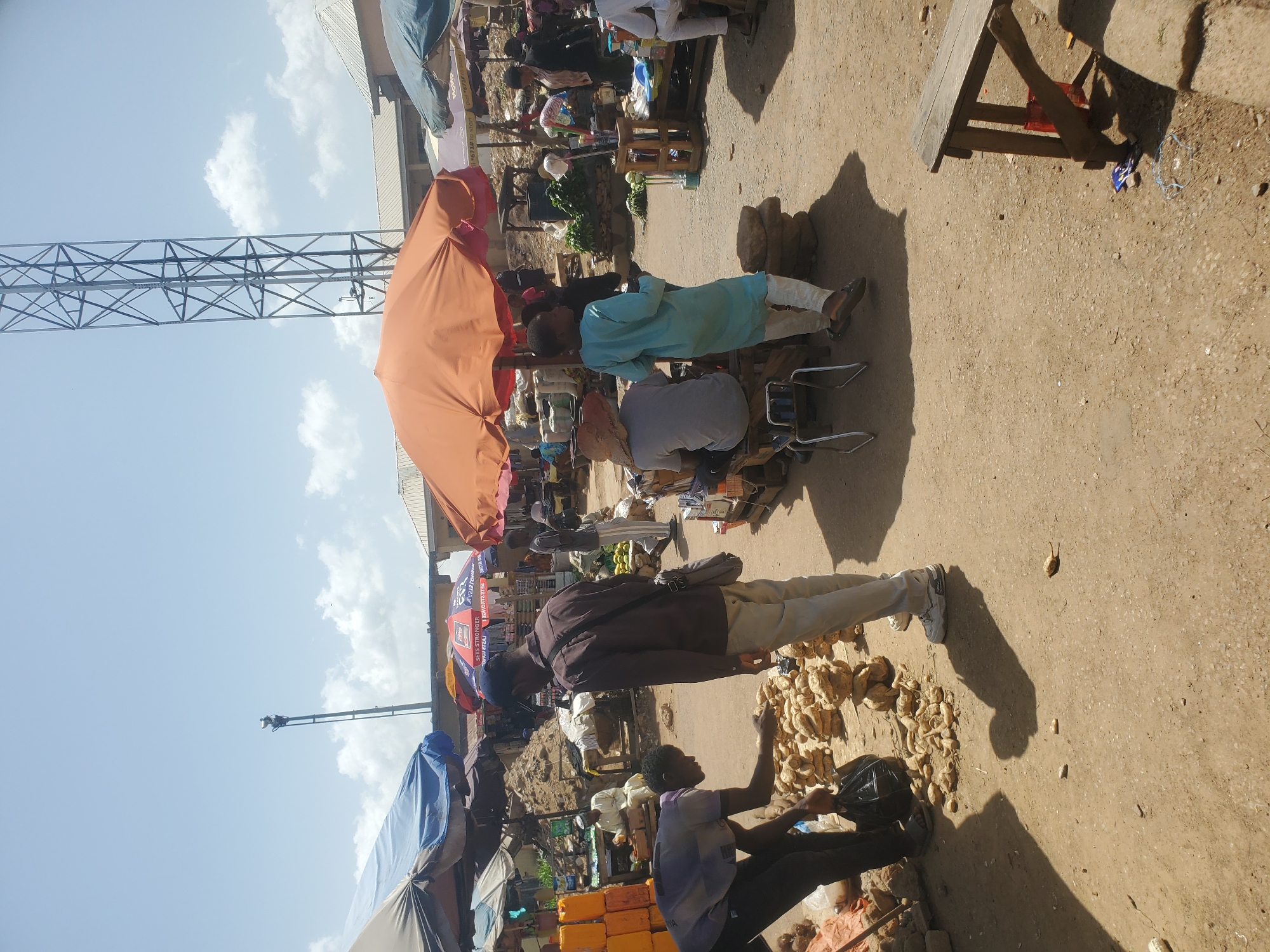 The image depicts a bustling outdoor market scene in Jos, Nigeria, under a clear, cloudy sky. In the foreground, a vendor, wearing a purple shirt and dark pants, is handing a black plastic bag to another person. Piles of root vegetables, likely yams or cassava, are stacked on the ground in front of them, with more scattered around. The person receiving the bag wears light-colored pants and white sneakers, with a dark bag slung over their shoulder.
Behind them, an orange umbrella shades a group of people. One individual, wearing a light blue robe and pants, stands facing away from the camera, while another sits on a stool wearing a wide-brimmed hat and a light gray garment. A tall metal tower, possibly a communication tower, dominates the background, with buildings and stalls visible further in the distance. Several other stalls are set up, indicated by umbrellas and piles of goods. The ground is unpaved and dusty, typical of an open-air market. The overall impression is one of active commerce and daily life.
