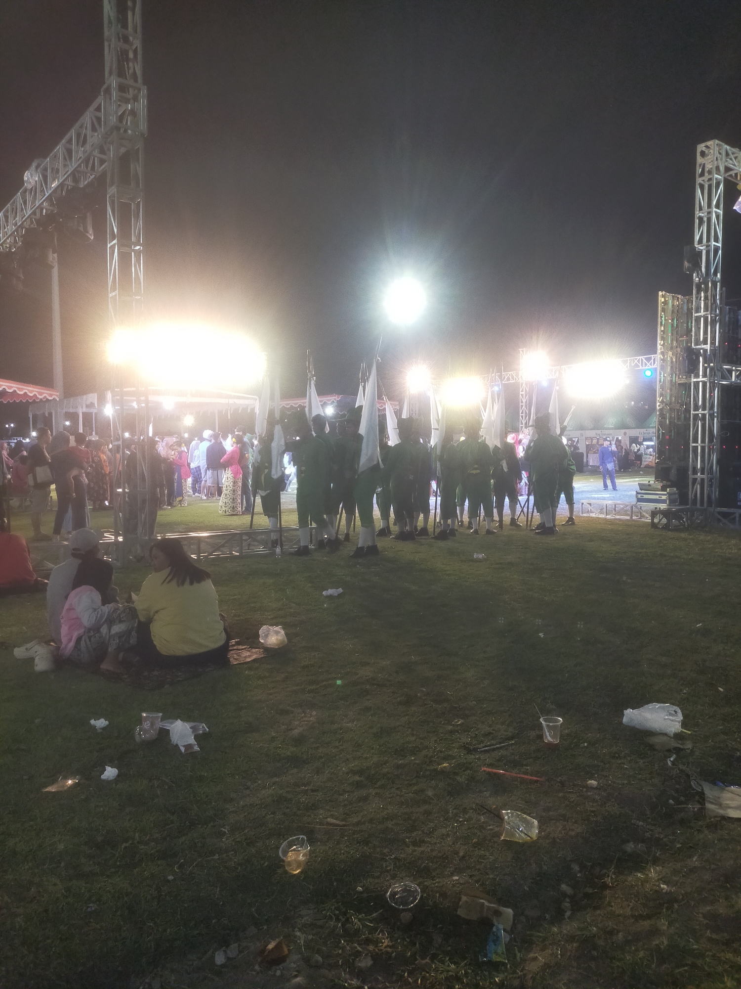 The image depicts an outdoor evening event, possibly a parade or ceremony, in the city of Bantul, Indonesia. In the foreground, several people are seated on the grass, facing away from the camera. The middle ground features a group of individuals in uniform, holding flags. Bright lights illuminate the scene from behind, suggesting a stage or performance area. In the background, more people are gathered, and structures that appear to be stages or scaffolding are visible. The ground is covered in grass and scattered with litter, including plastic cups and bags. The overall atmosphere is that of a public gathering at night, with an emphasis on a formal procession or display.